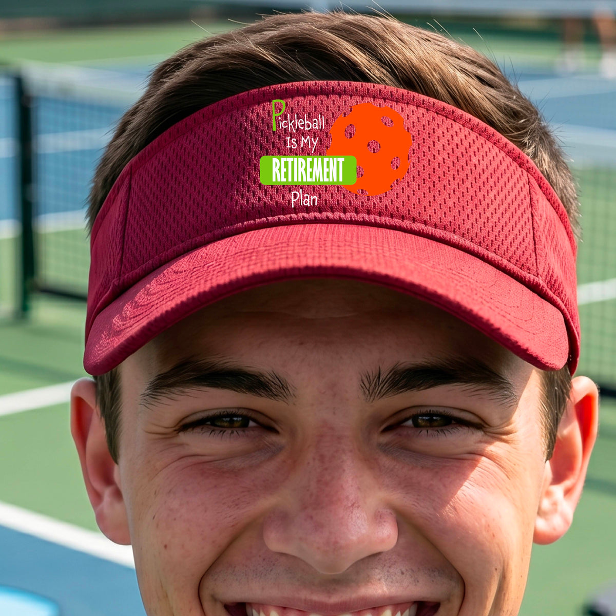 A close-up of a red visor with a custom graphic that reads 'Pickleball is my Retirement Plan' with a green and orange design, set against a blurred outdoor background.