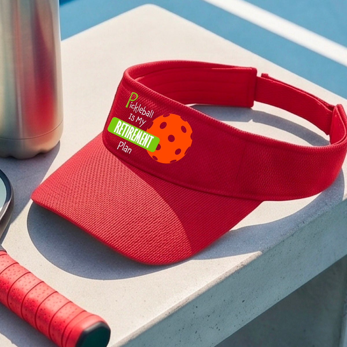 A close-up of a red visor with a custom graphic that reads 'Pickleball is my Retirement Plan' with a green and orange design, set against a blurred outdoor background.
