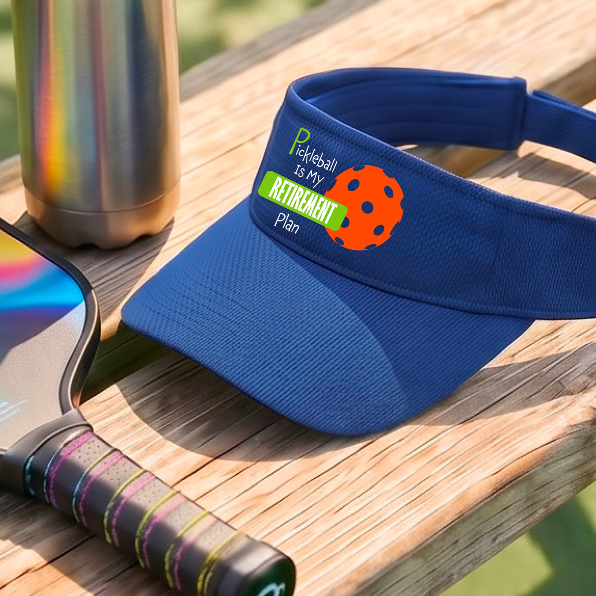 A close-up of a royal blue visor with a custom graphic that reads 'Pickleball is my Retirement Plan' with a green and orange design, set against a blurred outdoor background.