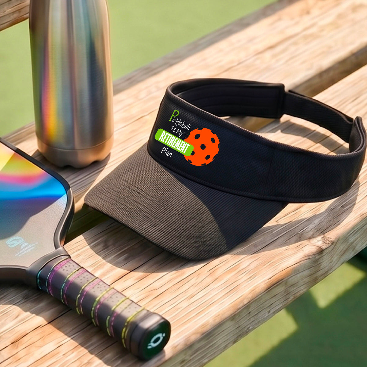 A close-up of a black visor with a custom graphic that reads 'Pickleball is my Retirement Plan' with a green and orange design, set against a blurred outdoor background.