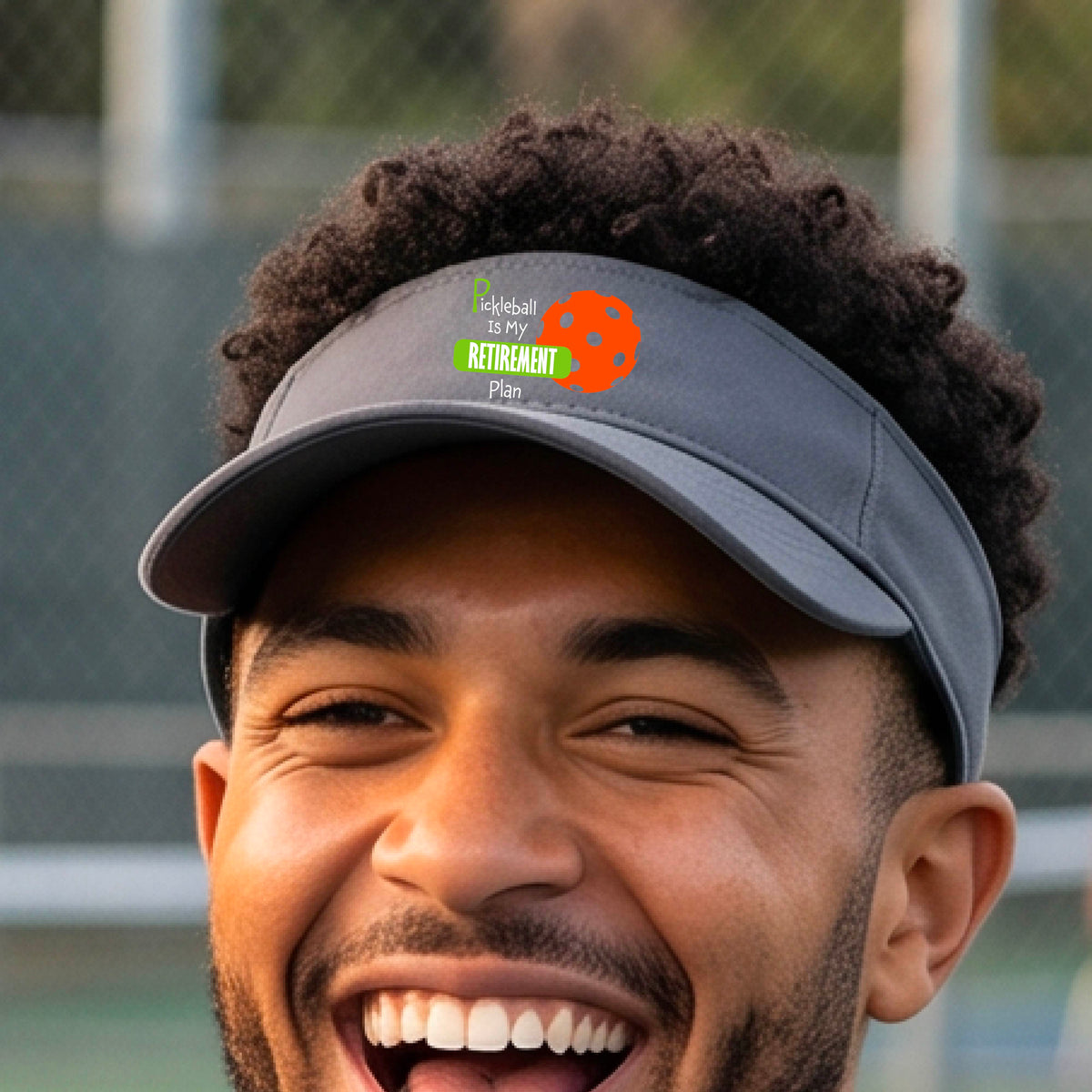 A close-up of a dark gray visor with a custom graphic that reads 'Pickleball is my Retirement Plan' with a green and orange design, set against a blurred outdoor background.