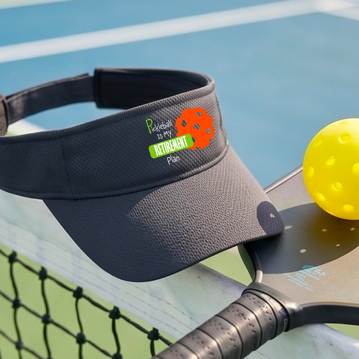 A close-up of a dark gray visor with a custom graphic that reads 'Pickleball is my Retirement Plan' with a green and orange design, set against a blurred outdoor background.