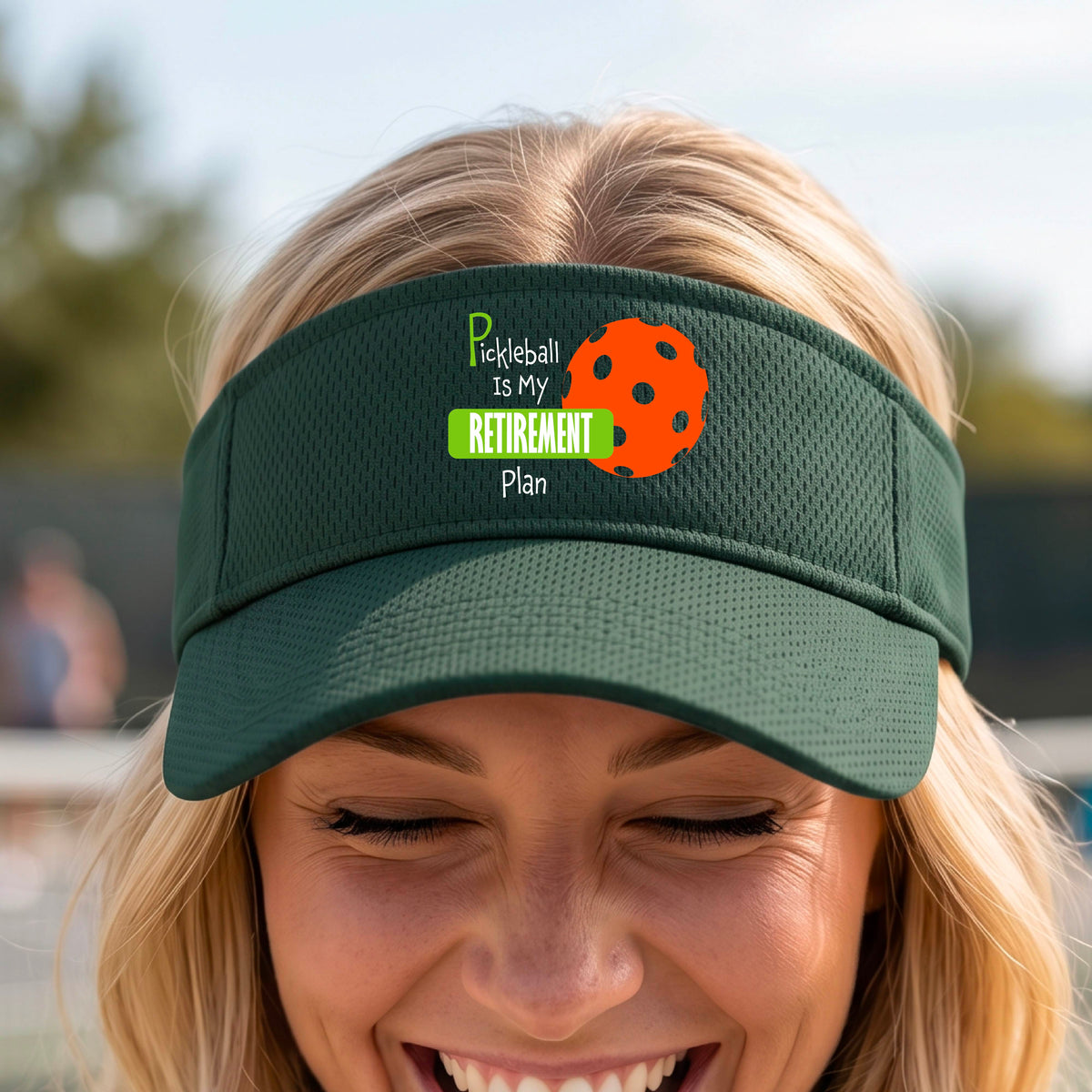 A close-up of a green visor with a custom graphic that reads 'Pickleball is my Retirement Plan' with a green and orange design, set against a blurred outdoor background.