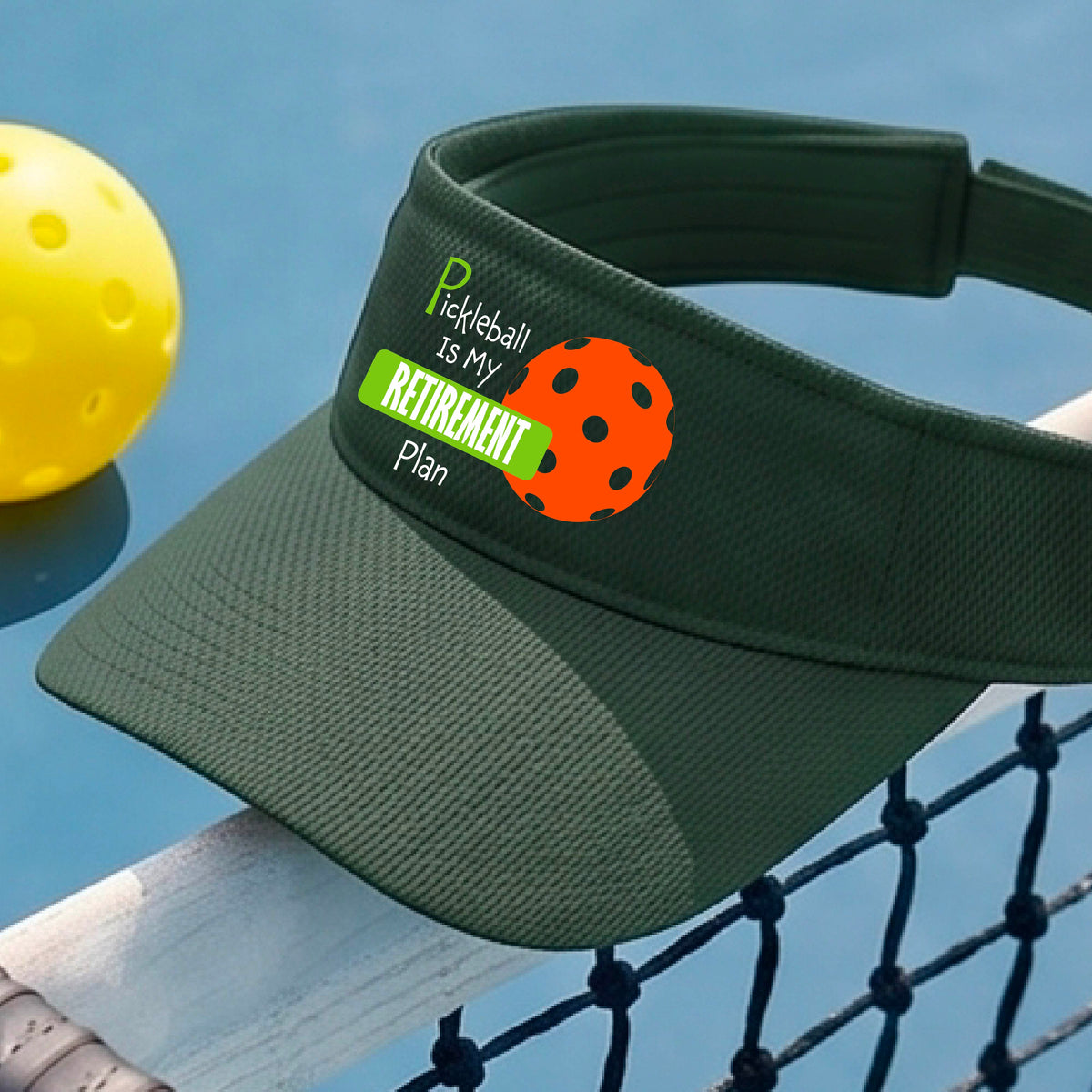 A close-up of a green visor with a custom graphic that reads 'Pickleball is my Retirement Plan' with a green and orange design, set against a blurred outdoor background.