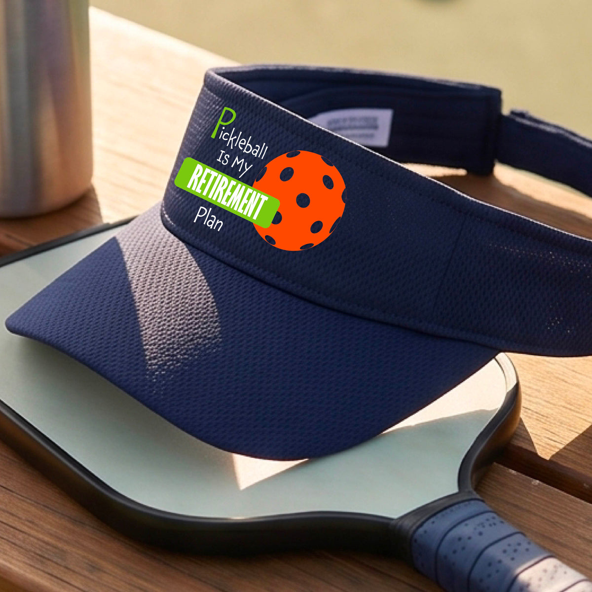 A close-up of a navy blue visor with a custom graphic that reads 'Pickleball is my Retirement Plan' with a green and orange design, set against a blurred outdoor background.
