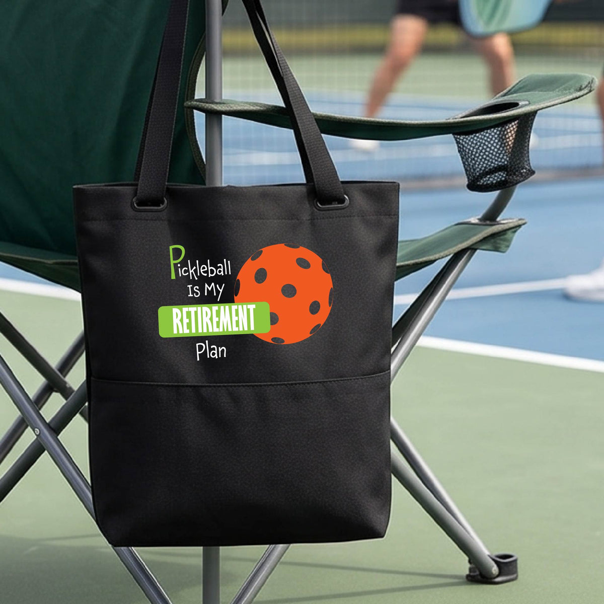 A close up of a medium size black tote bag on a pickleball court, with a graphic of the words “Pickleball is my Retirement Plan” in green and white with an orange pickleball illustration in the background.
