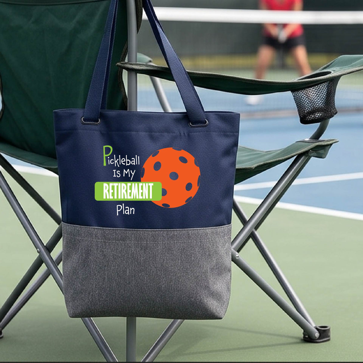A close up of a medium size navy blue and grey tote bag on a pickleball court, with a graphic of the words “Pickleball is my Retirement Plan” in green and white with an orange pickleball illustration in the background.