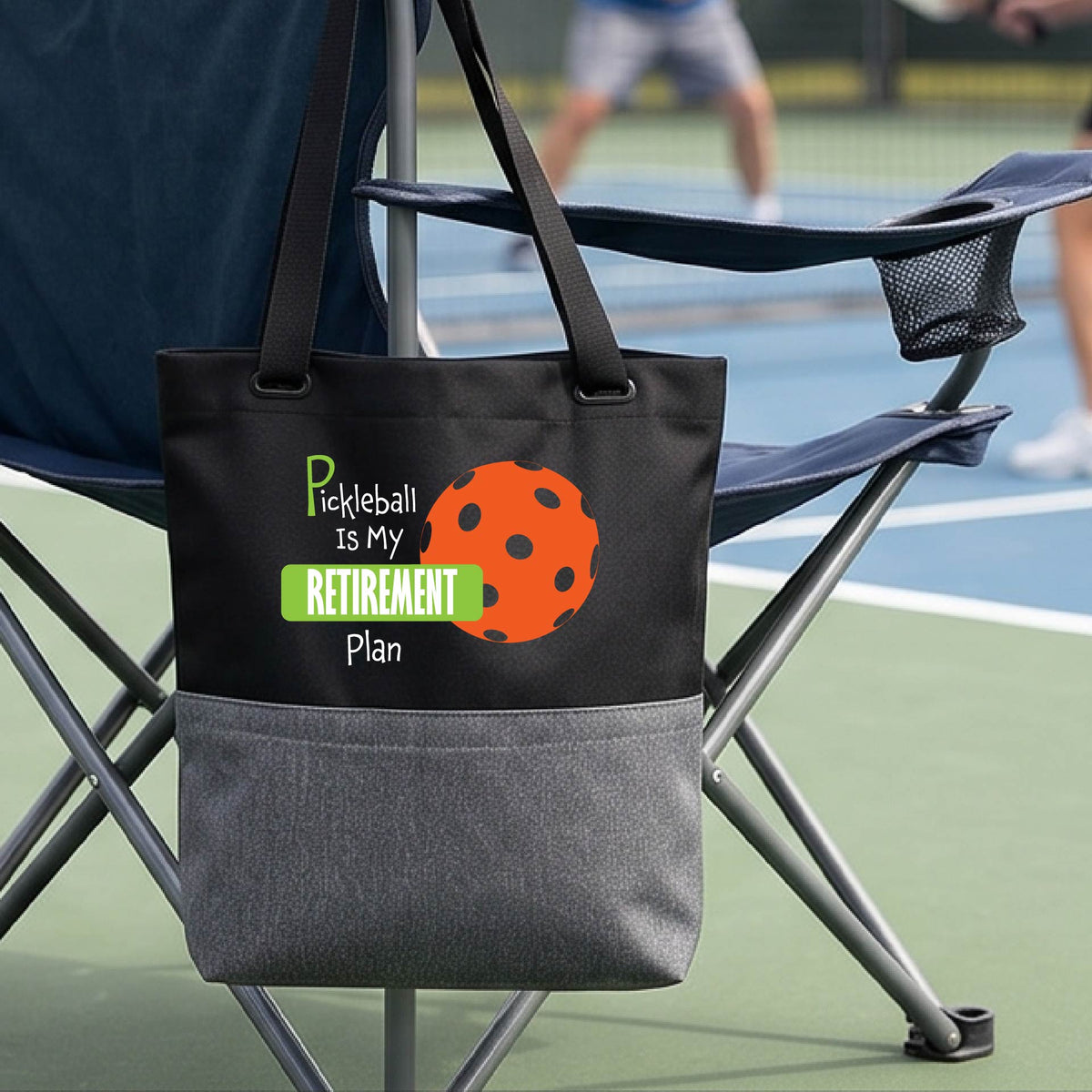 A close up of a medium size black and grey tote bag on a pickleball court, with a graphic of the words “Pickleball is my Retirement Plan” in green and white with an orange pickleball illustration in the background.