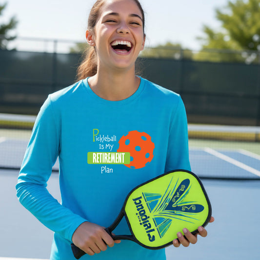 A young person on a pickleball court, wearing a bright blue long-sleeve athletic t-shirt, with the words “Pickleball is my Retirement Plan” in green and white with an orange pickleball illustration in the background.