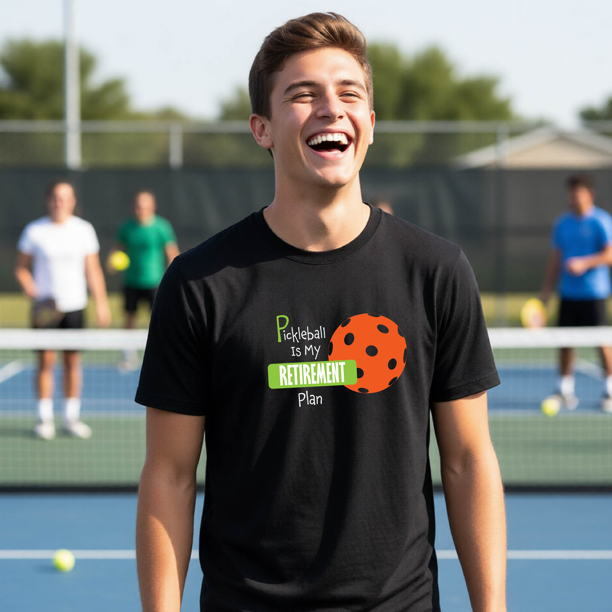 A young person laughing on an outdoor tennis court wearing a black t-shirt with a graphic that reads 'Pickleball is my RETIREMENT Plan' alongside an orange pickleball illustration.