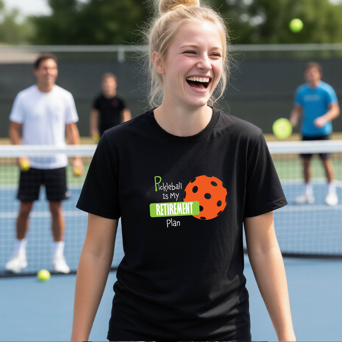 A young person laughing on an outdoor tennis court wearing a black t-shirt with a graphic that reads 'Pickleball is my RETIREMENT Plan' alongside an orange pickleball illustration.