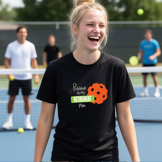 A young person laughing on an outdoor tennis court wearing a black t-shirt with a graphic that reads 'Pickleball is my RETIREMENT Plan' alongside an orange pickleball illustration.