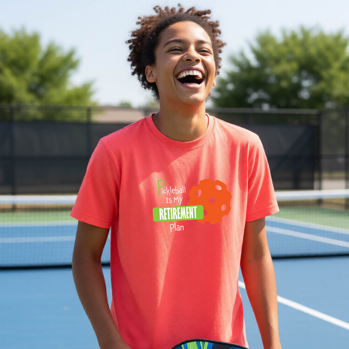 A young person laughing on an outdoor tennis court wearing a coral pink t-shirt with a graphic that reads 'Pickleball is my RETIREMENT Plan' alongside an orange pickleball illustration.