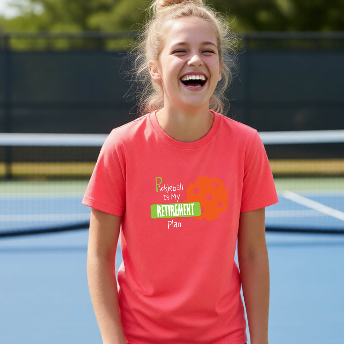 A young person laughing on an outdoor tennis court wearing a coral pink t-shirt with a graphic that reads 'Pickleball is my RETIREMENT Plan' alongside an orange pickleball illustration.