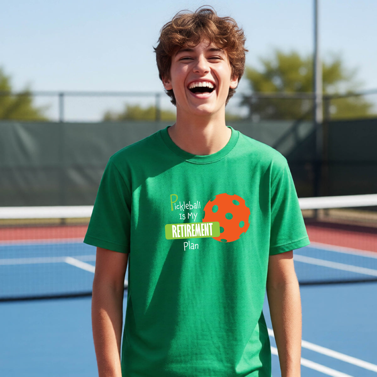 A young person laughing on an outdoor tennis court wearing a green t-shirt with a graphic that reads 'Pickleball is my RETIREMENT Plan' alongside an orange pickleball illustration.