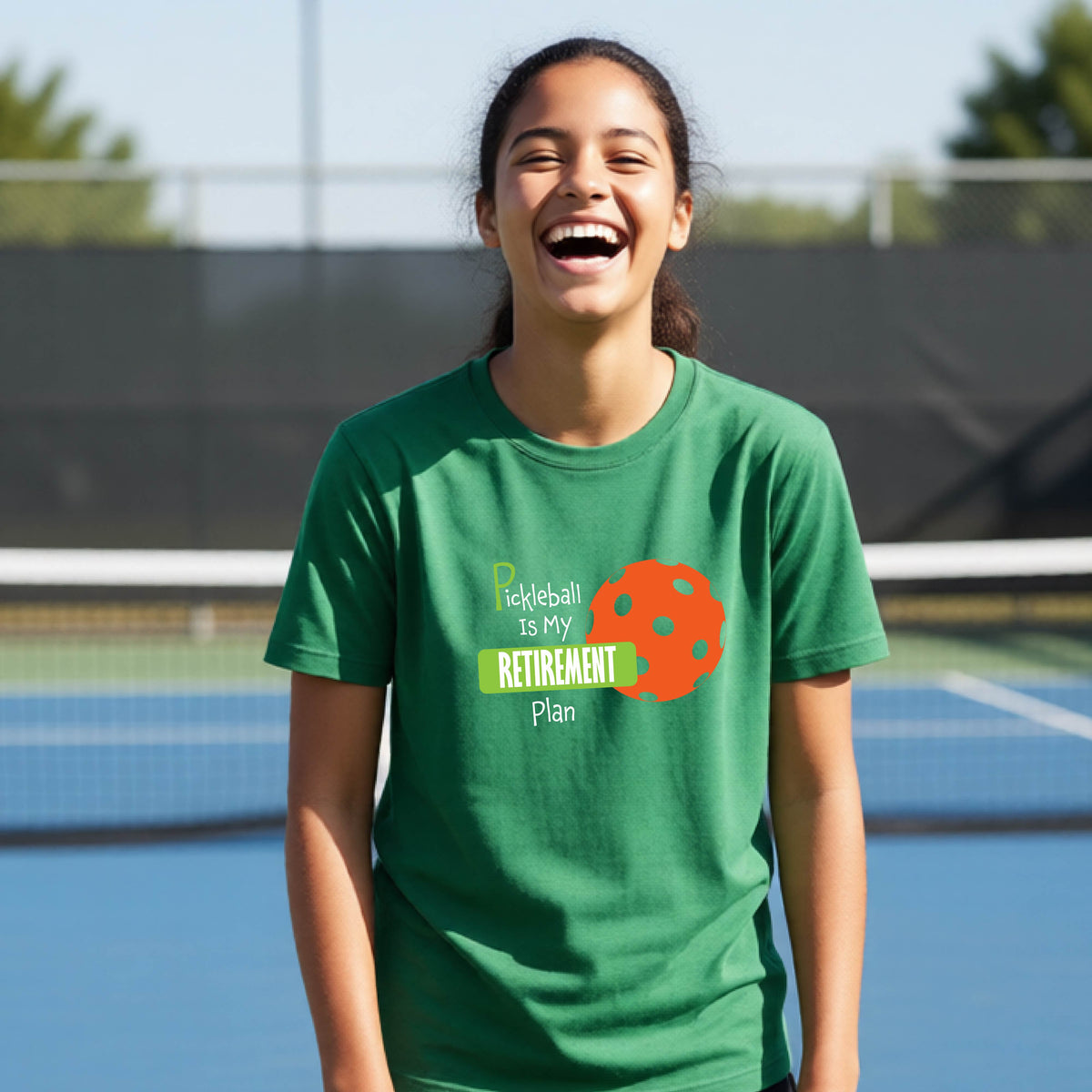 A young person laughing on an outdoor tennis court wearing a green t-shirt with a graphic that reads 'Pickleball is my RETIREMENT Plan' alongside an orange pickleball illustration.