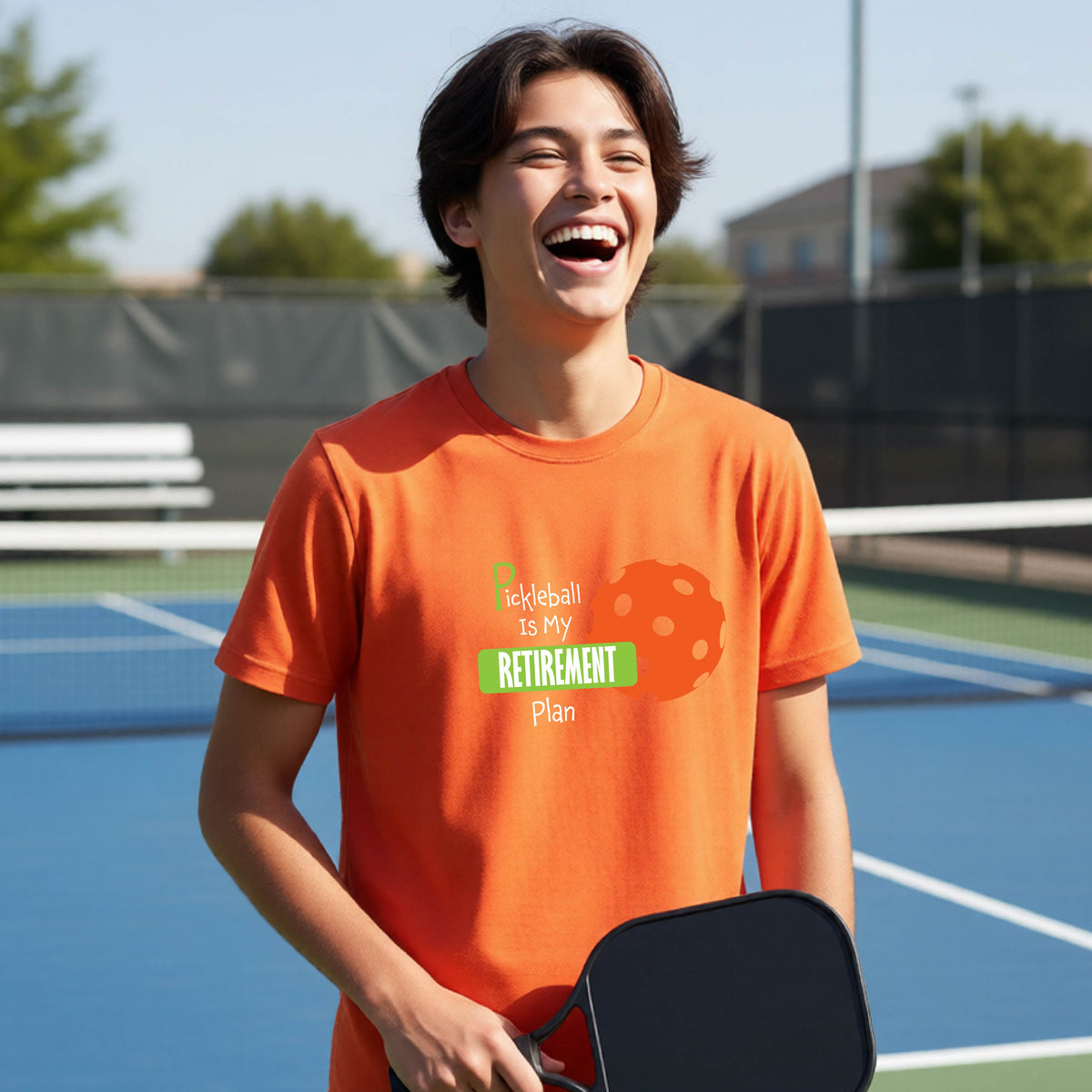 A young person laughing on an outdoor tennis court wearing an orange t-shirt with a graphic that reads 'Pickleball is my RETIREMENT Plan' alongside an orange pickleball illustration.