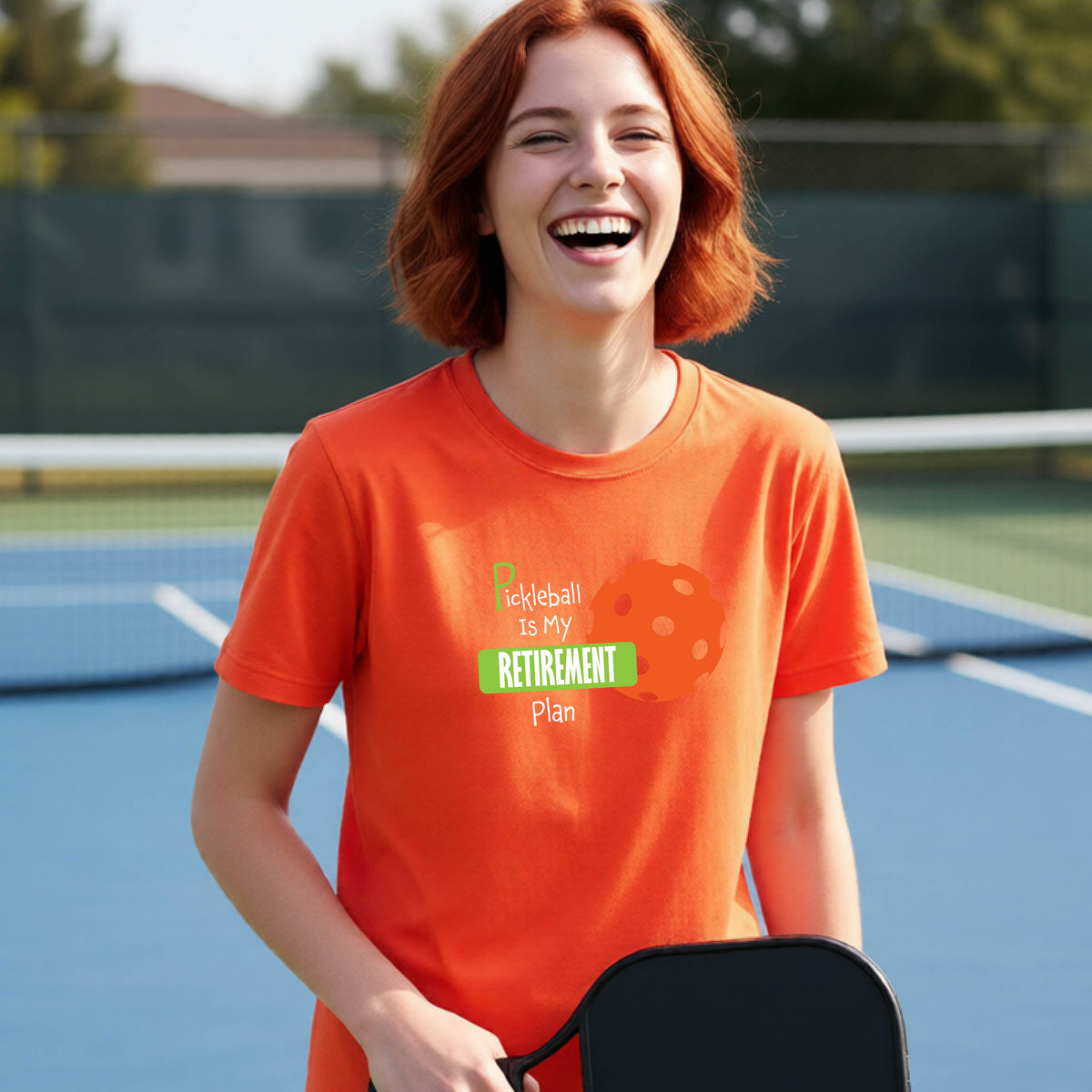A young person laughing on an outdoor tennis court wearing an orange t-shirt with a graphic that reads 'Pickleball is my RETIREMENT Plan' alongside an orange pickleball illustration.