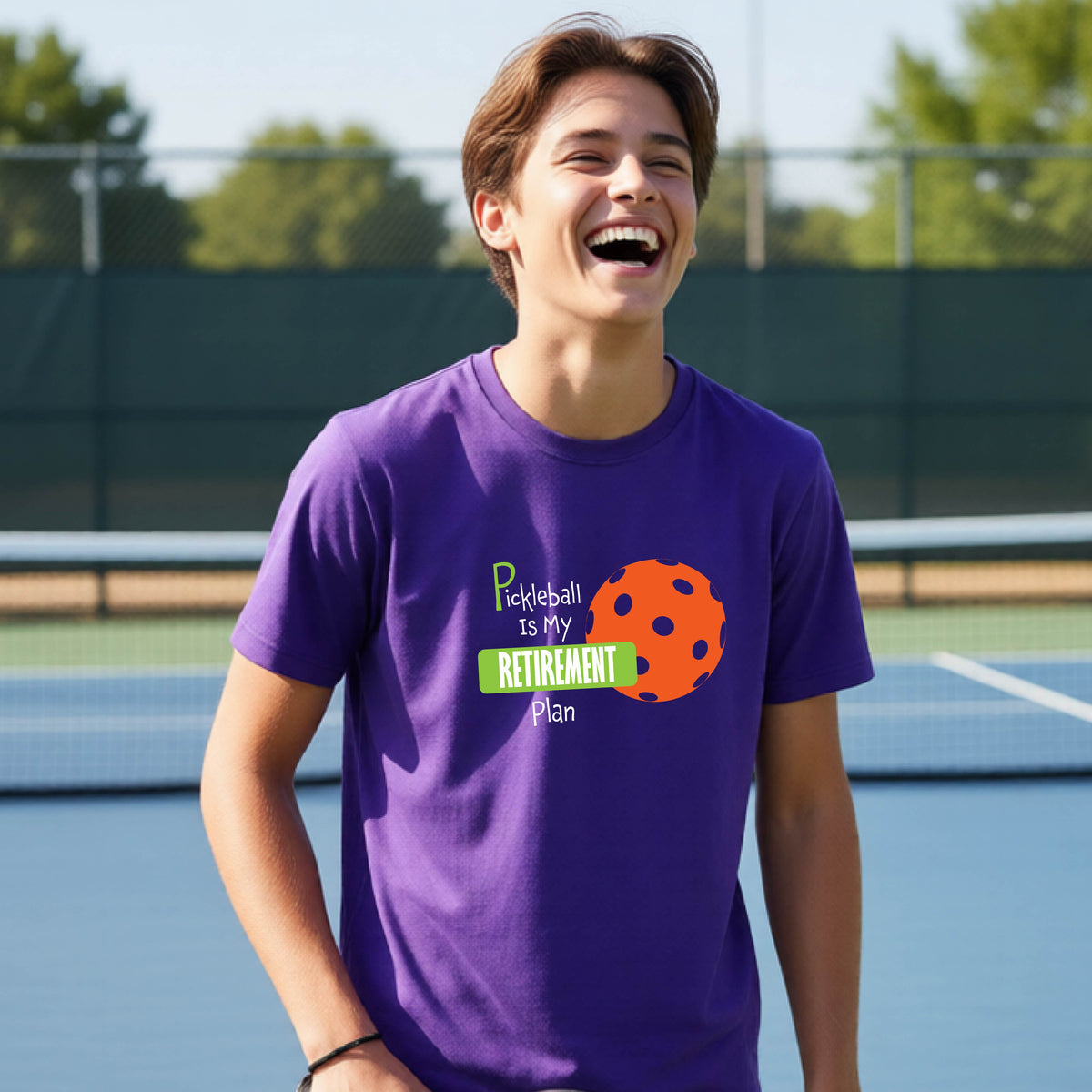 A young person laughing on an outdoor tennis court wearing a purple t-shirt with a graphic that reads 'Pickleball is my RETIREMENT Plan' alongside an orange pickleball illustration.