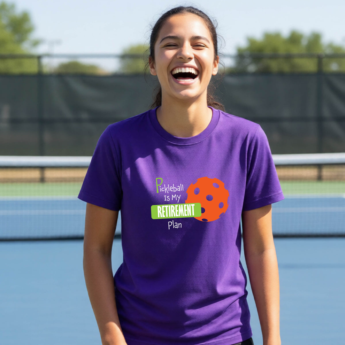 A young person laughing on an outdoor tennis court wearing a purple t-shirt with a graphic that reads 'Pickleball is my RETIREMENT Plan' alongside an orange pickleball illustration.