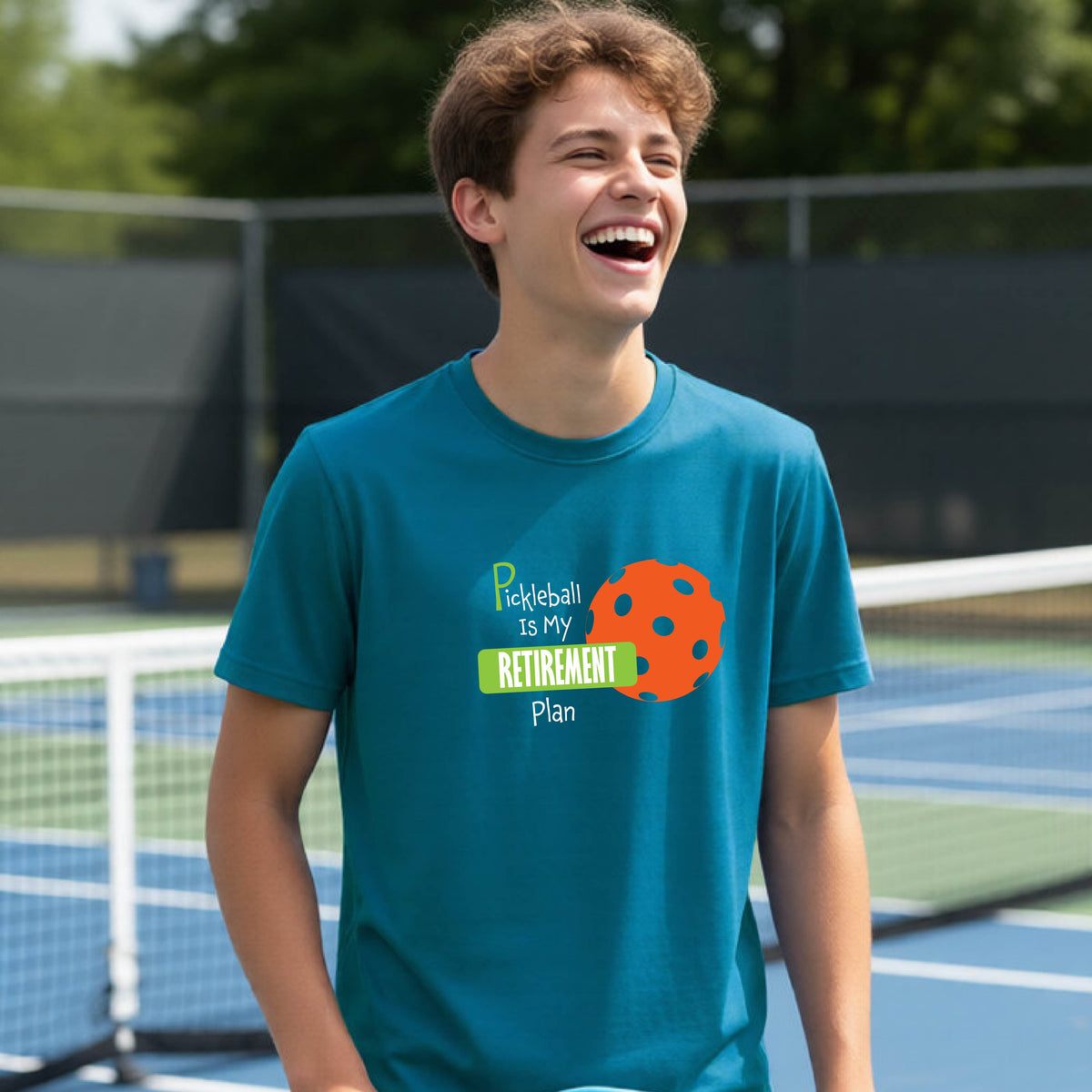 A young person laughing on an outdoor tennis court wearing a teal t-shirt with a graphic that reads 'Pickleball is my RETIREMENT Plan' alongside an orange pickleball illustration.