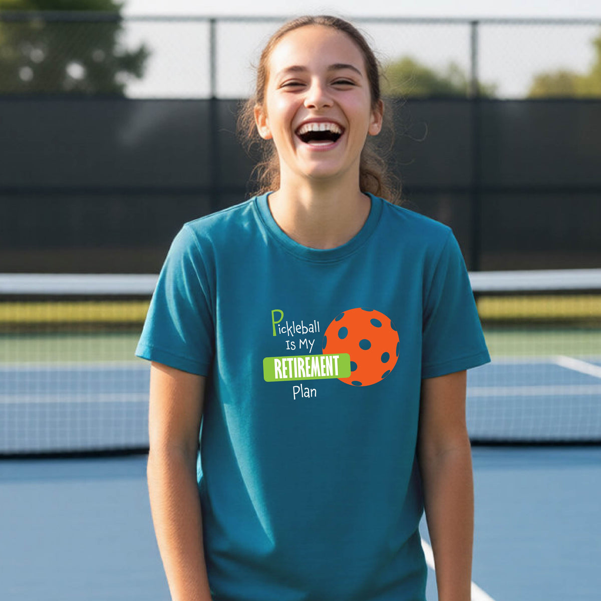 A young person laughing on an outdoor tennis court wearing a teal t-shirt with a graphic that reads 'Pickleball is my RETIREMENT Plan' alongside an orange pickleball illustration.