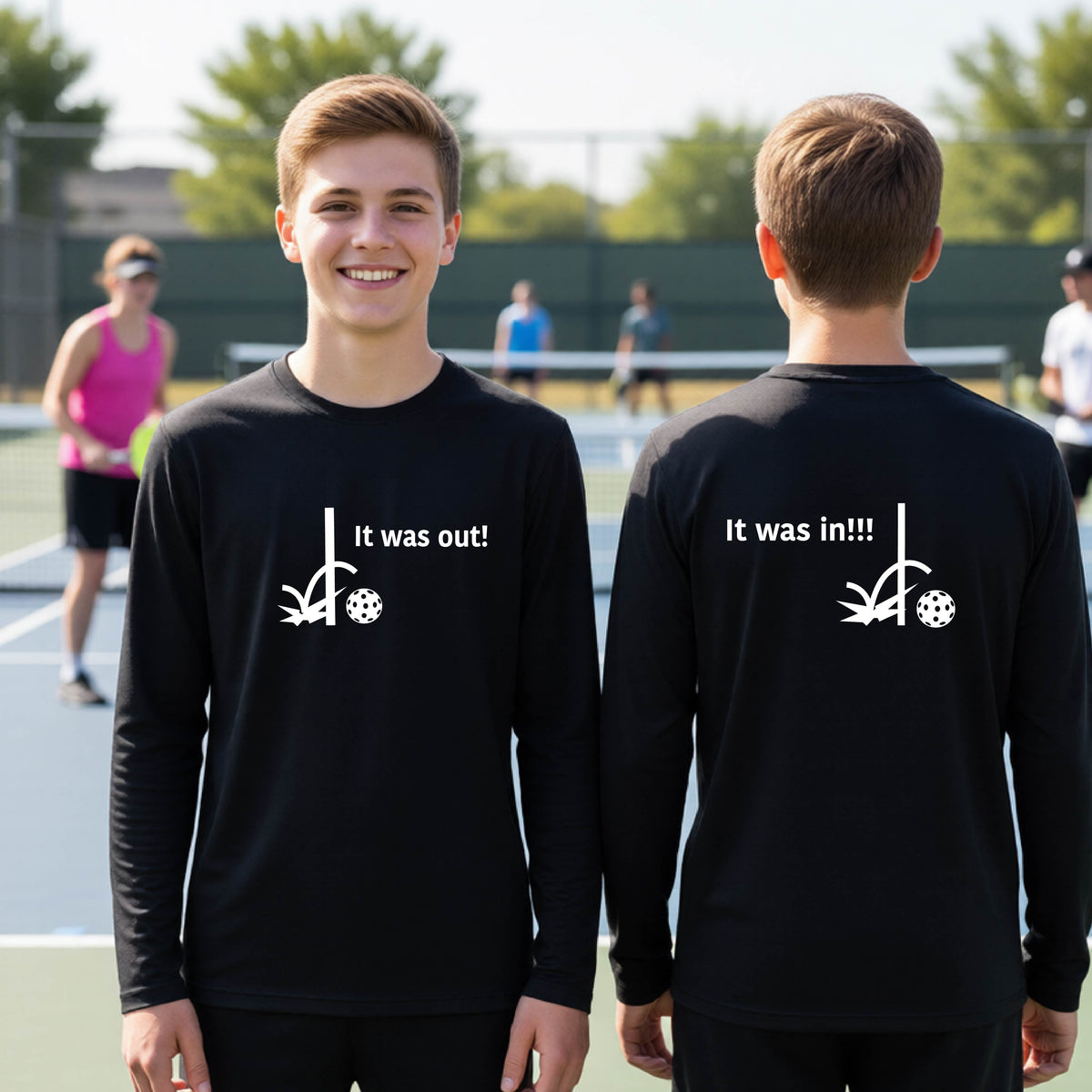 Two youths showing a black  long-sleeve t-shirt with a white design of ‘IT WAS OUT!’ and a colorful pickleball on the front, and the back design with a white ‘IT WAS IN!!!’ and a colorful pickleball, standing on a pickleball court.