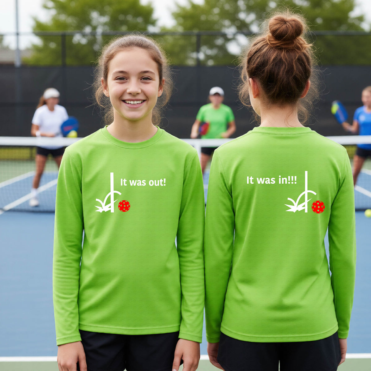 Two youths showing a lime green long-sleeve t-shirt with a white design of ‘IT WAS OUT!’ and a colorful pickleball on the front, and the back design with a white ‘IT WAS IN!!!’ and a colorful pickleball, standing on a pickleball court.