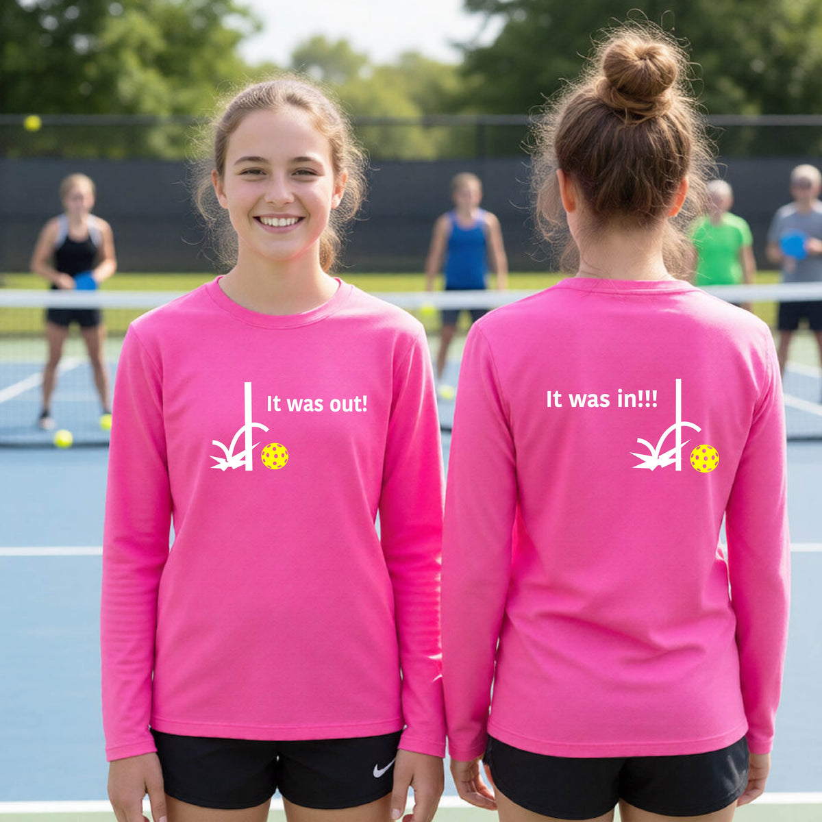 Two youths showing a pink long-sleeve t-shirt with a white design of ‘IT WAS OUT!’ and a colorful pickleball on the front, and the back design with a white ‘IT WAS IN!!!’ and a colorful pickleball, standing on a pickleball court.