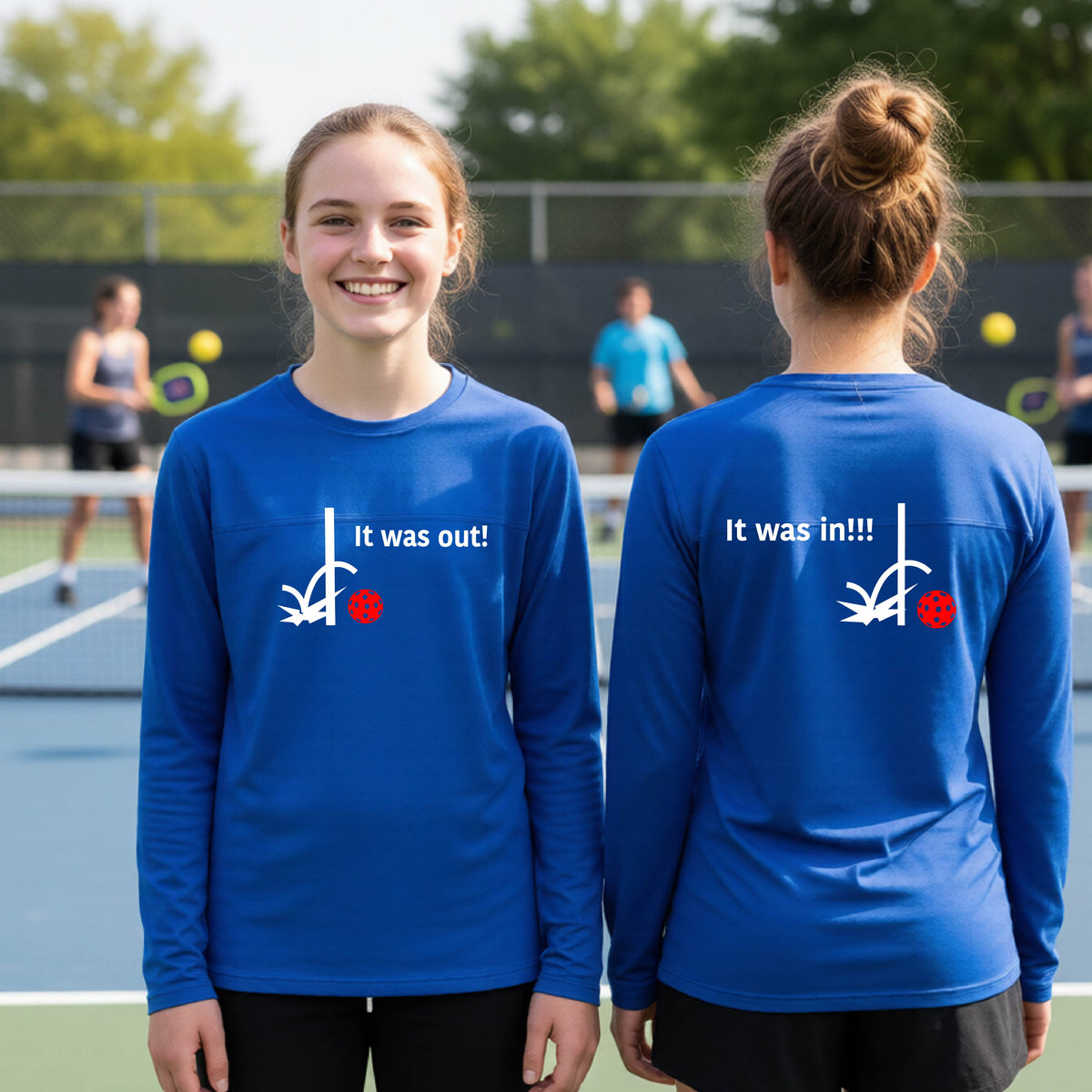 Two youths showing a royal blue long-sleeve t-shirt with a white design of ‘IT WAS OUT!’ and a colorful pickleball on the front, and the back design with a white ‘IT WAS IN!!!’ and a colorful pickleball, standing on a pickleball court.