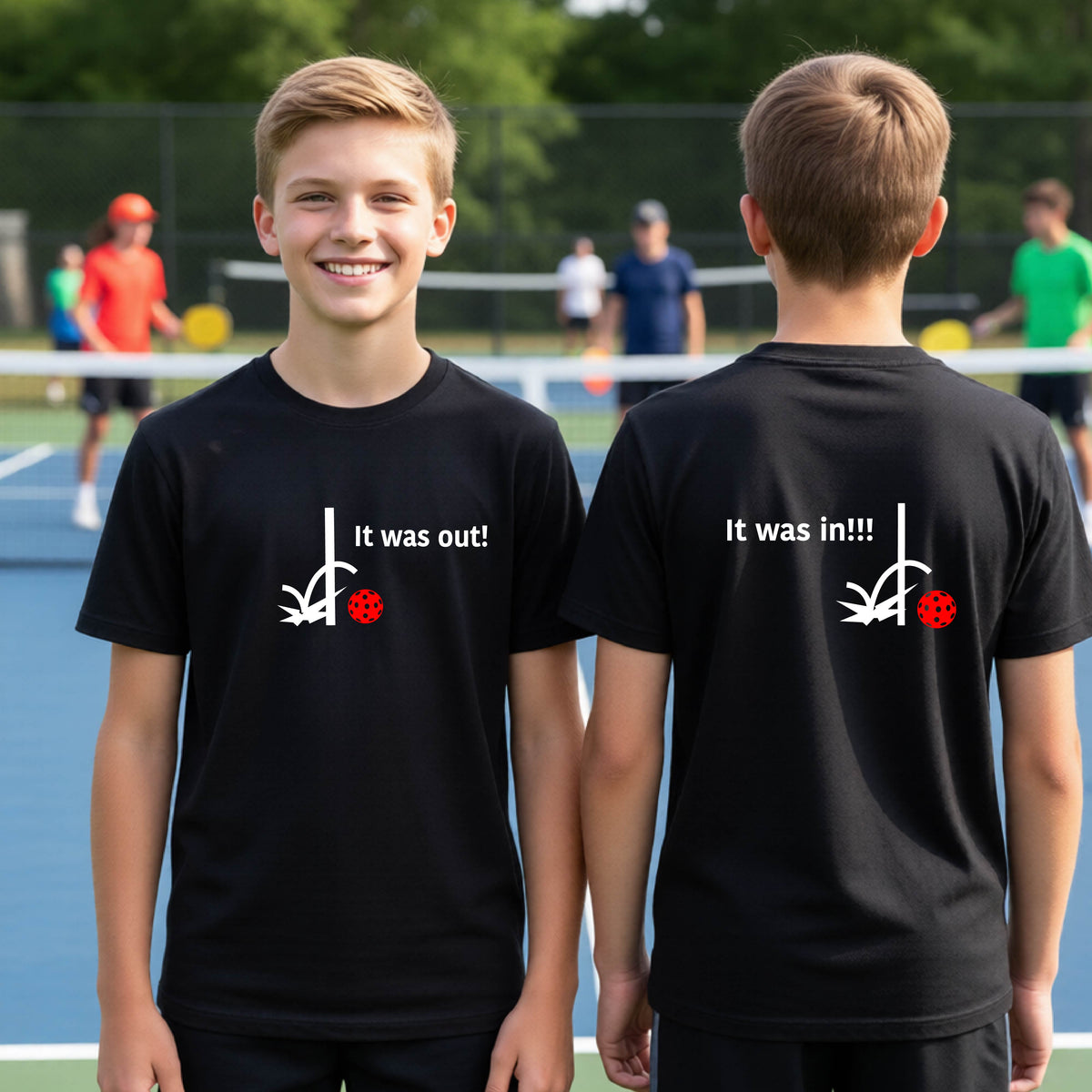 Two youths showing a black short-sleeve t-shirt with a white design of ‘IT WAS OUT!’ and a colorful pickleball on the front, and the back design with a white ‘IT WAS IN!!!’ and a colorful pickleball, standing on a pickleball court.