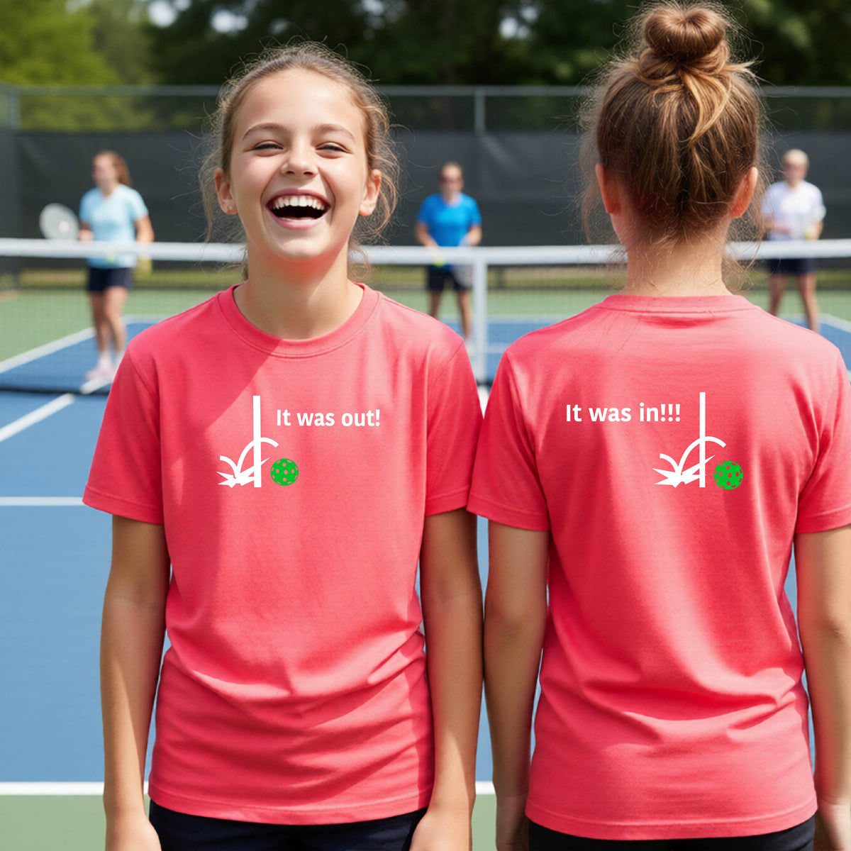 Two youths showing a coral pink short-sleeve t-shirt with a white design of ‘IT WAS OUT!’ and a colorful pickleball on the front, and the back design with a white ‘IT WAS IN!!!’ and a colorful pickleball, standing on a pickleball court.