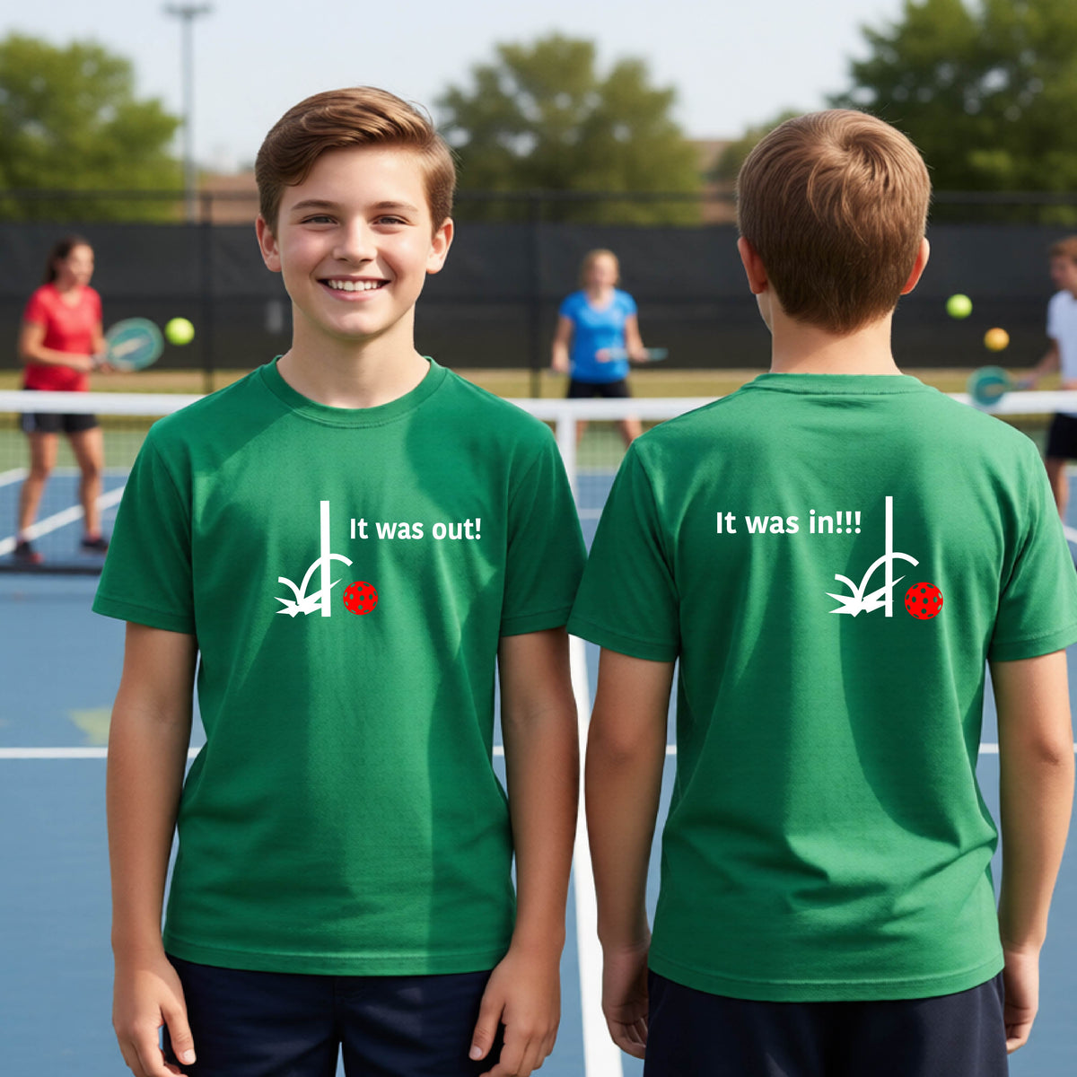 Two youths showing a green short-sleeve t-shirt with a white design of ‘IT WAS OUT!’ and a colorful pickleball on the front, and the back design with a white ‘IT WAS IN!!!’ and a colorful pickleball, standing on a pickleball court.