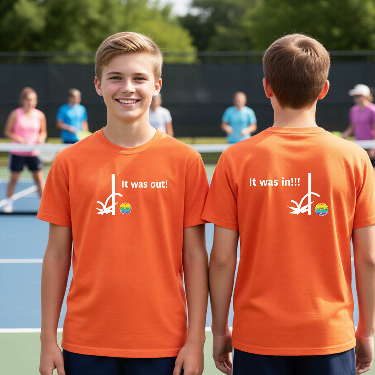 Two youths showing an orange short-sleeve t-shirt with a white design of ‘IT WAS OUT!’ and a colorful pickleball on the front, and the back design with a white ‘IT WAS IN!!!’ and a colorful pickleball, standing on a pickleball court.