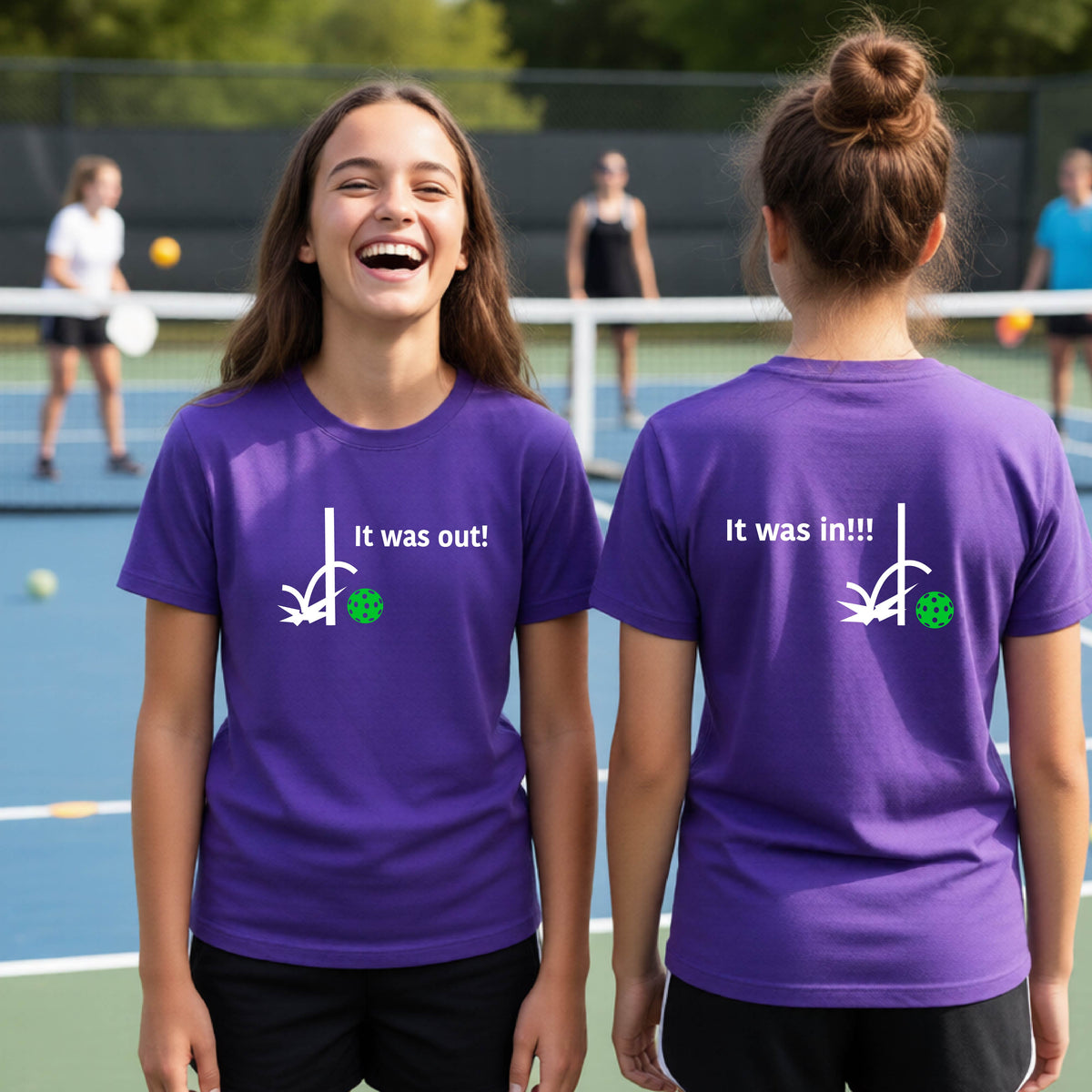 Two youths showing a purple short-sleeve t-shirt with a white design of ‘IT WAS OUT!’ and a colorful pickleball on the front, and the back design with a white ‘IT WAS IN!!!’ and a colorful pickleball, standing on a pickleball court.
