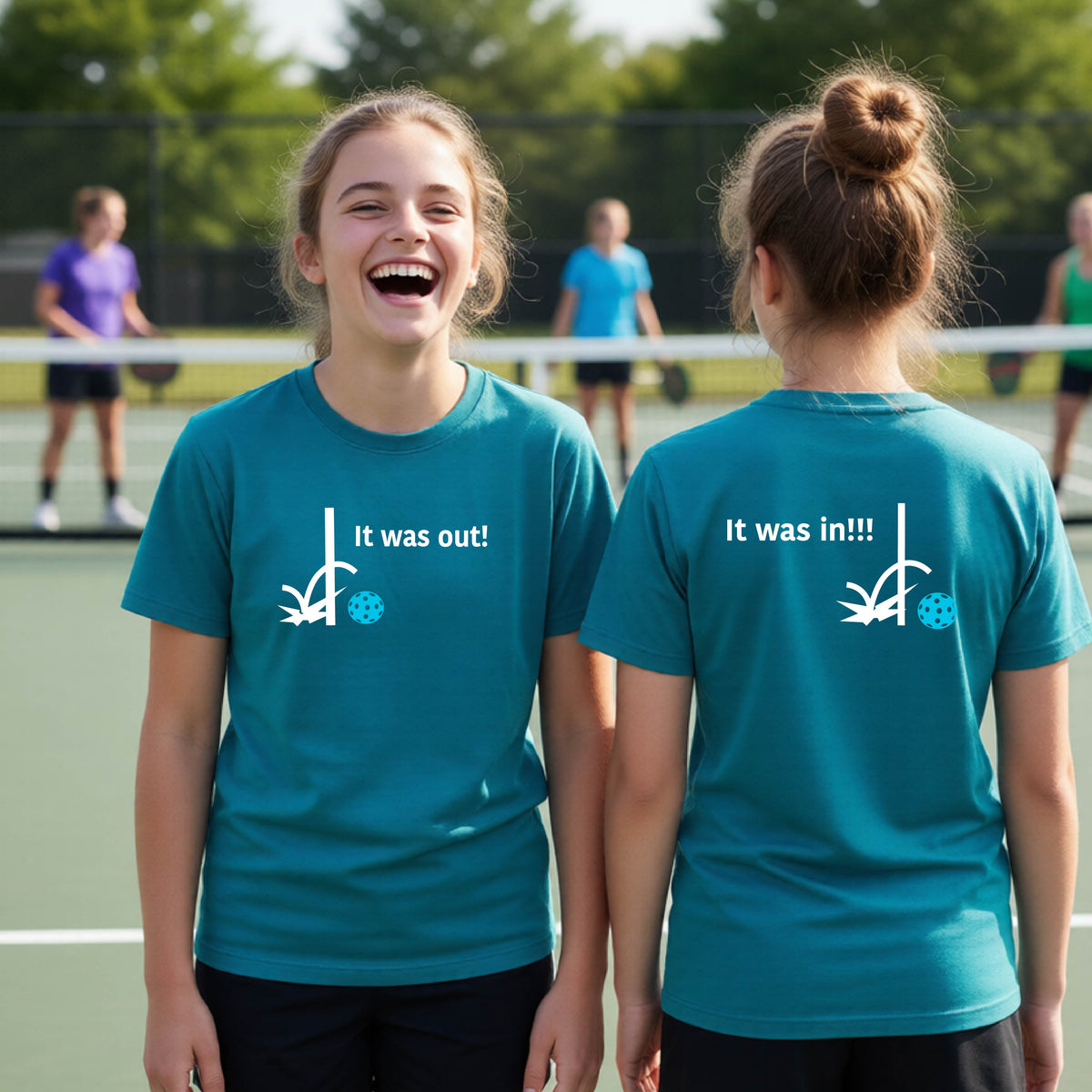 Two youths showing a teal short-sleeve t-shirt with a white design of ‘IT WAS OUT!’ and a colorful pickleball on the front, and the back design with a white ‘IT WAS IN!!!’ and a colorful pickleball, standing on a pickleball court.