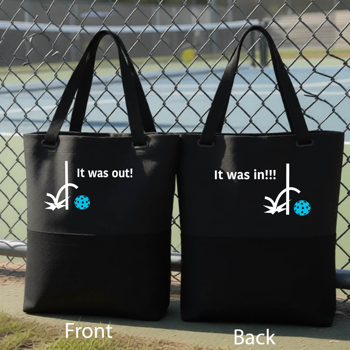 Two black tote bags with a white design of ‘IT WAS OUT!’ and a colorful pickleball on the front, and the back design with a white ‘IT WAS IN!!!’ and a colorful pickleball, standing on a pickleball court.