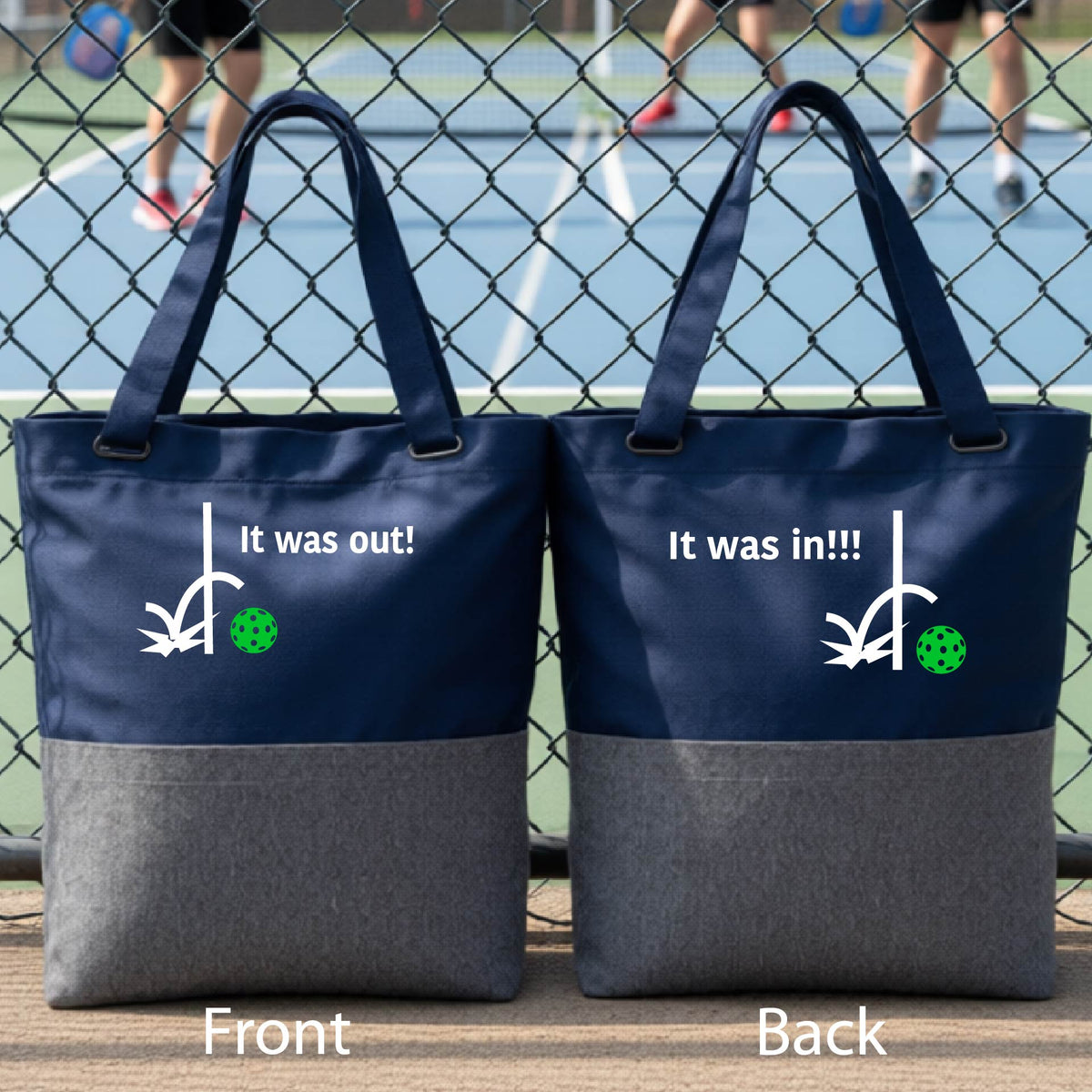 Two blue and gray tote bags with a white design of ‘IT WAS OUT!’ and a colorful pickleball on the front, and the back design with a white ‘IT WAS IN!!!’ and a colorful pickleball, standing on a pickleball court.