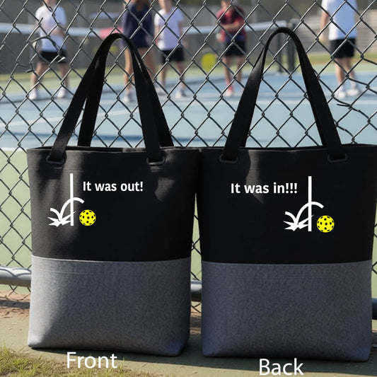 Two black and gray tote bags with a white design of ‘IT WAS OUT!’ and a colorful pickleball on the front, and the back design with a white ‘IT WAS IN!!!’ and a colorful pickleball, standing on a pickleball court.