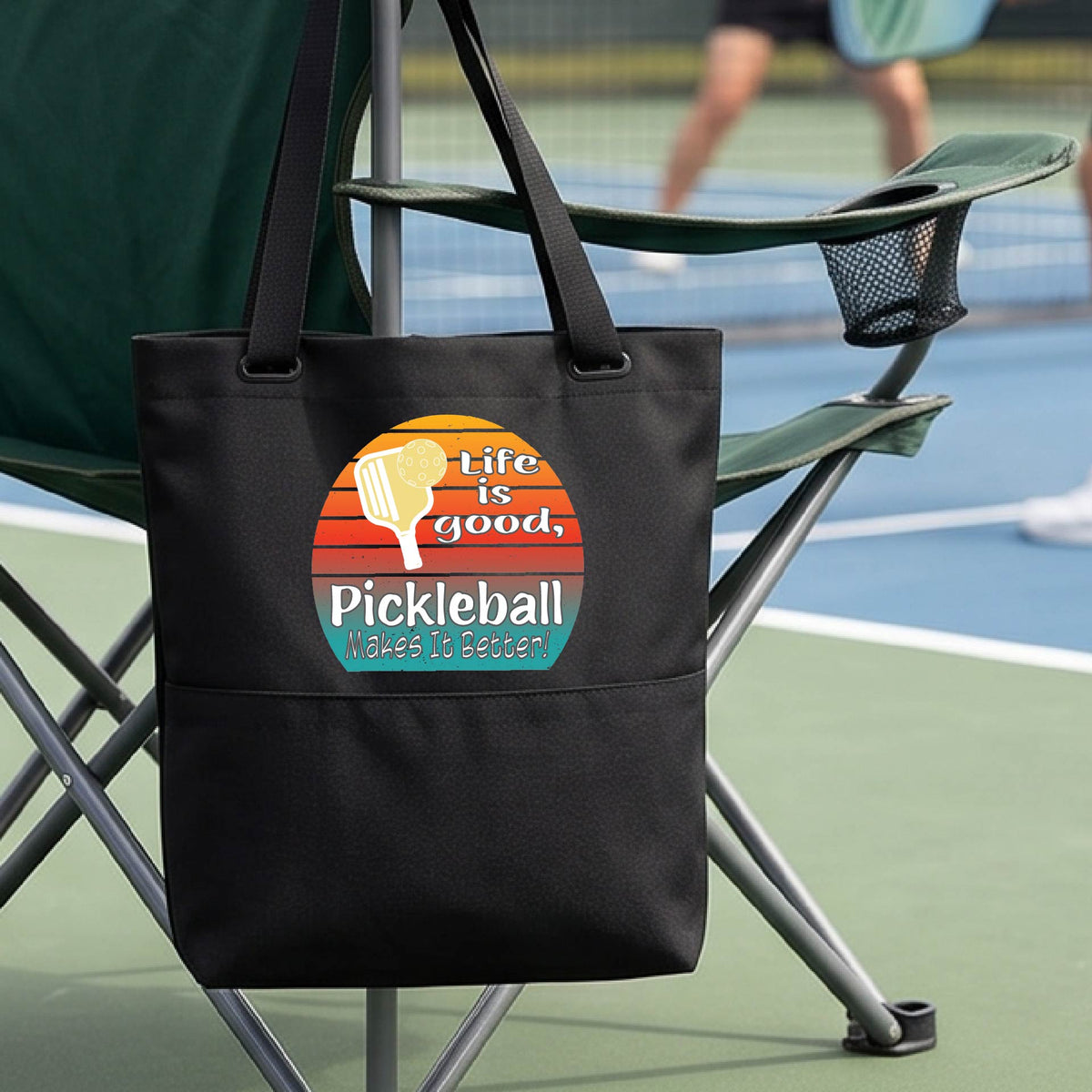 A black tote bag with a retro multi-colored circle with a pickleball paddle and ball, with white text that says 'LIFE IS GOOD PICKLEBALL MAKES IT BETTER', in front of a pickleball court.