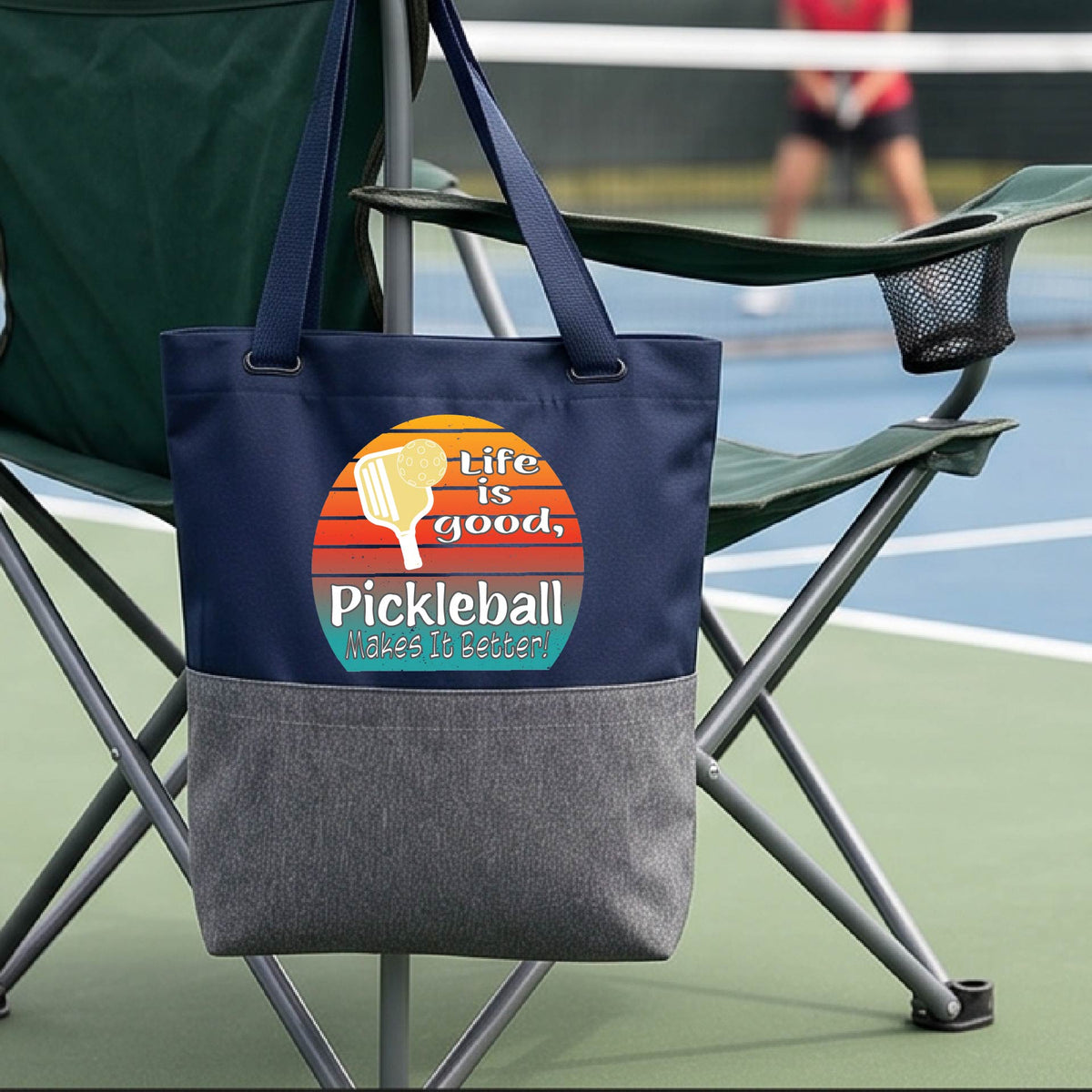 A blue and gray tote bag with a retro multi-colored circle with a pickleball paddle and ball, with white text that says 'LIFE IS GOOD PICKLEBALL MAKES IT BETTER', in front of a pickleball court.