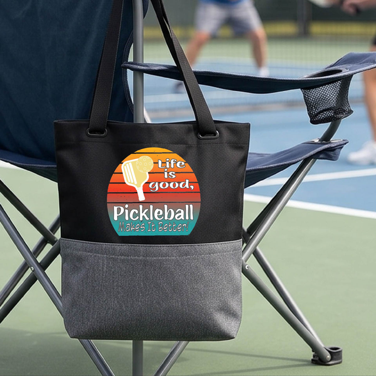 A black and gray tote bag with a retro multi-colored circle with a pickleball paddle and ball, with white text that says 'LIFE IS GOOD PICKLEBALL MAKES IT BETTER', in front of a pickleball court.