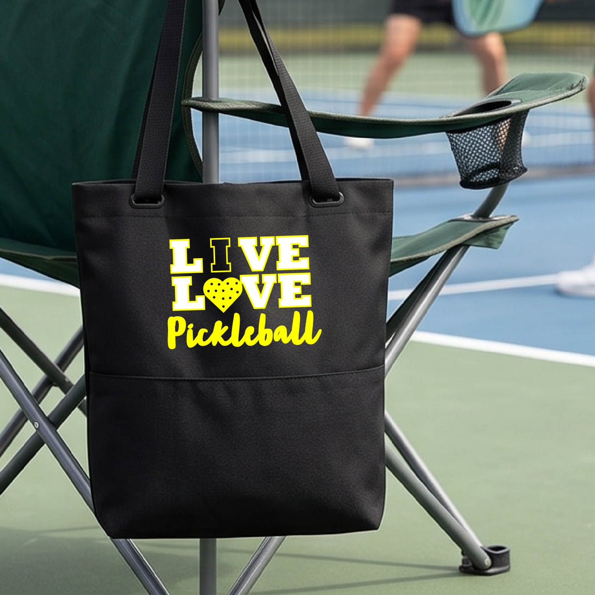 A black canvas tote bag with 'LIVE LOVE PICKLEBALL' printed in yellow and white on the front, hanging from a green folding chair on a pickleball court. A blurred player is visible in the background.