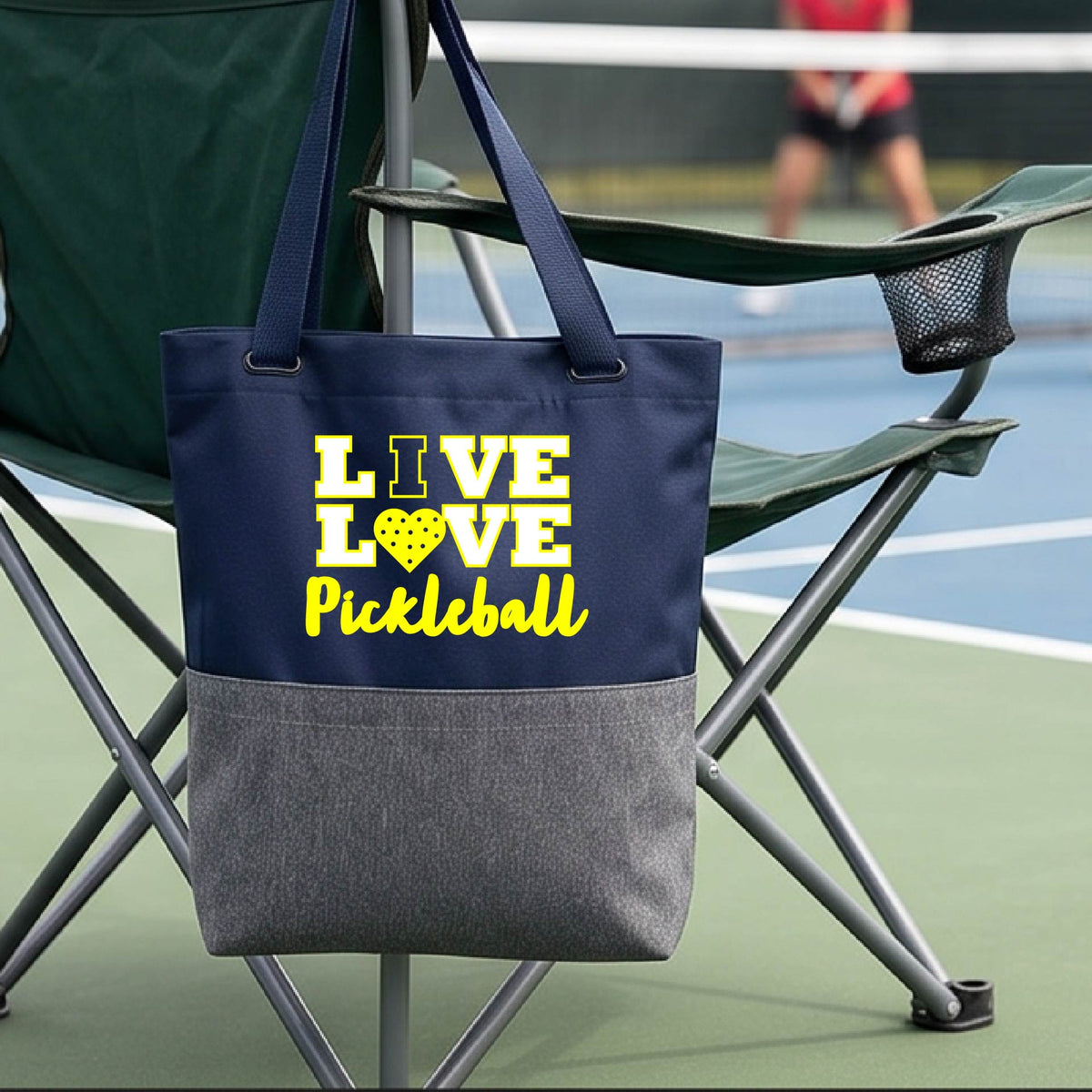 A navy blue and gray tote bag with yellow text reading 'LIVE LOVE Pickleball' hanging from a green folding chair on a tennis court, with a blurred player in the background.