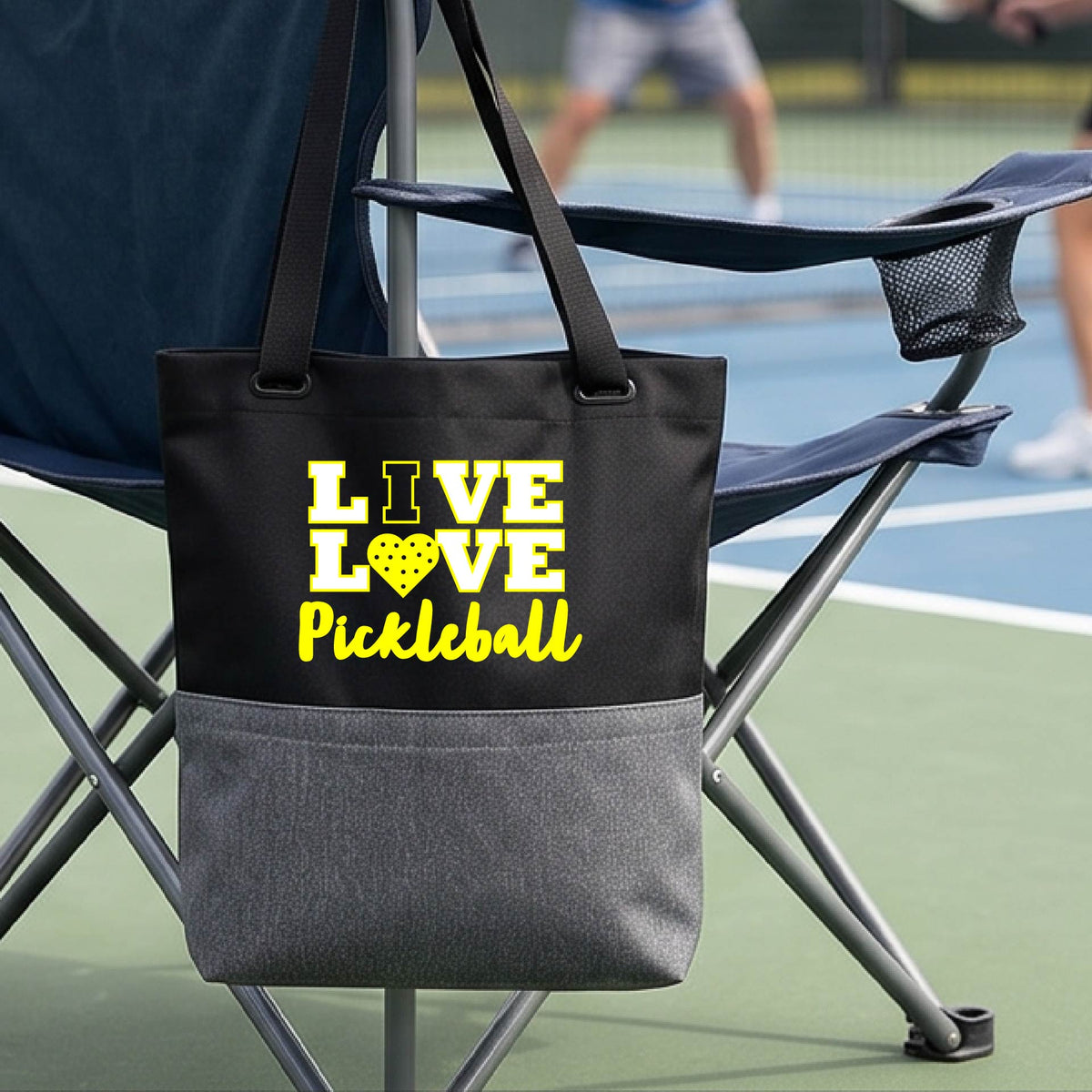 A black and gray tote bag with yellow text reading 'LIVE LOVE Pickleball' hanging from a folding chair on a pickleball court. The bag features a black upper section with the printed design and a heather gray lower pocket.