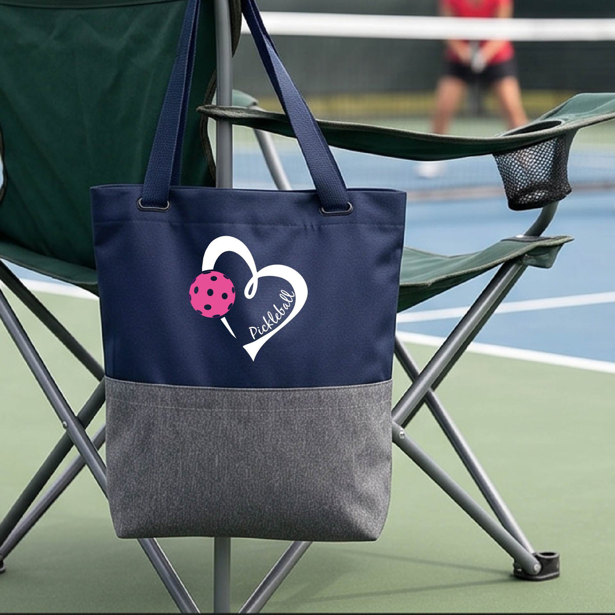 A blue and gray canvas tote bag with a white heart outline and a pink pickleball graphic, featuring the text 'Pickleball' on the front. The bag is hanging on a green folding chair on a tennis court, with a blurred player in the background.