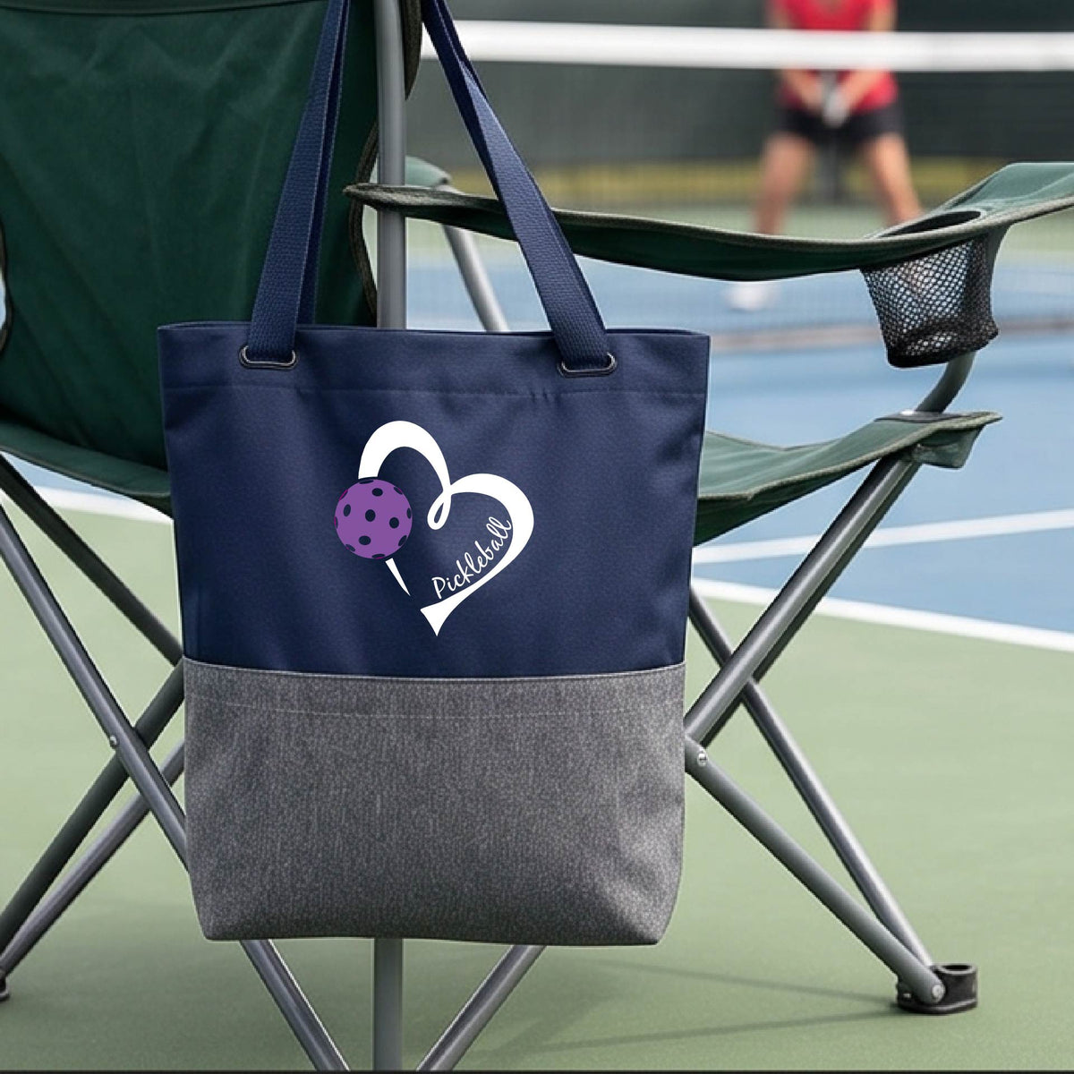 A blue and gray canvas tote bag with a white heart outline and a purple pickleball graphic, featuring the text 'Pickleball' on the front. The bag is hanging on a green folding chair on a tennis court, with a blurred player in the background.