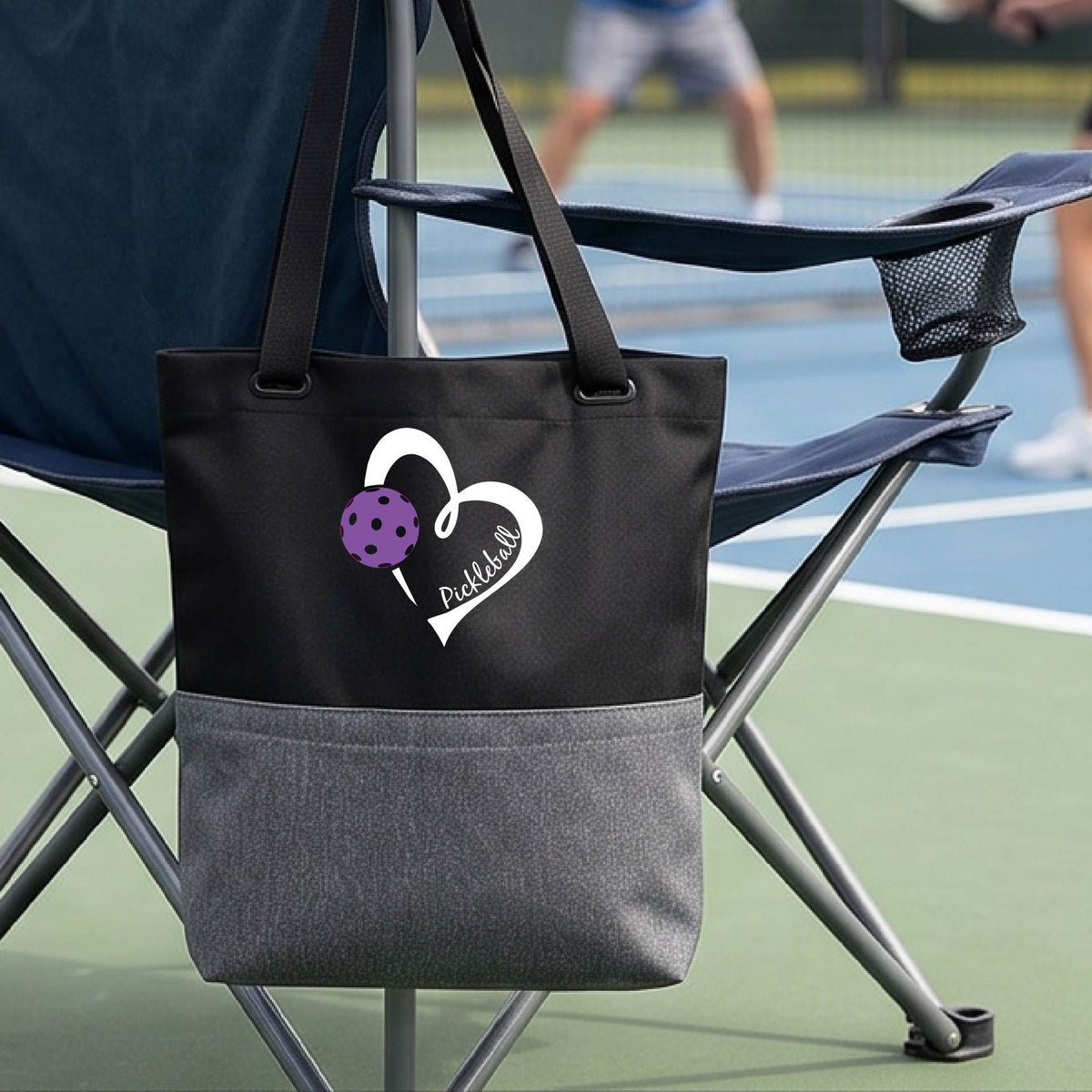 A black and gray canvas tote bag with a white heart outline and a purple pickleball graphic, featuring the text 'Pickleball' on the front. The bag is hanging on a green folding chair on a tennis court, with a blurred player in the background.