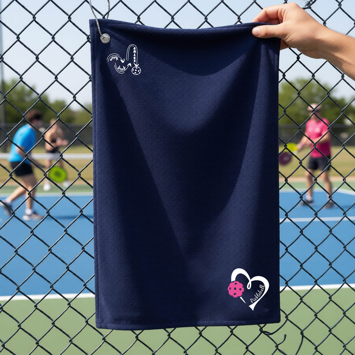 A navy blue terry cloth towel held up by a hand in front of a chain-link fence and outdoor pickleball court. The towel has a white stylized 'DINK DINK SMASH' logo near the top left and a white heart outline with a pink pickleball graphic near the bottom right.