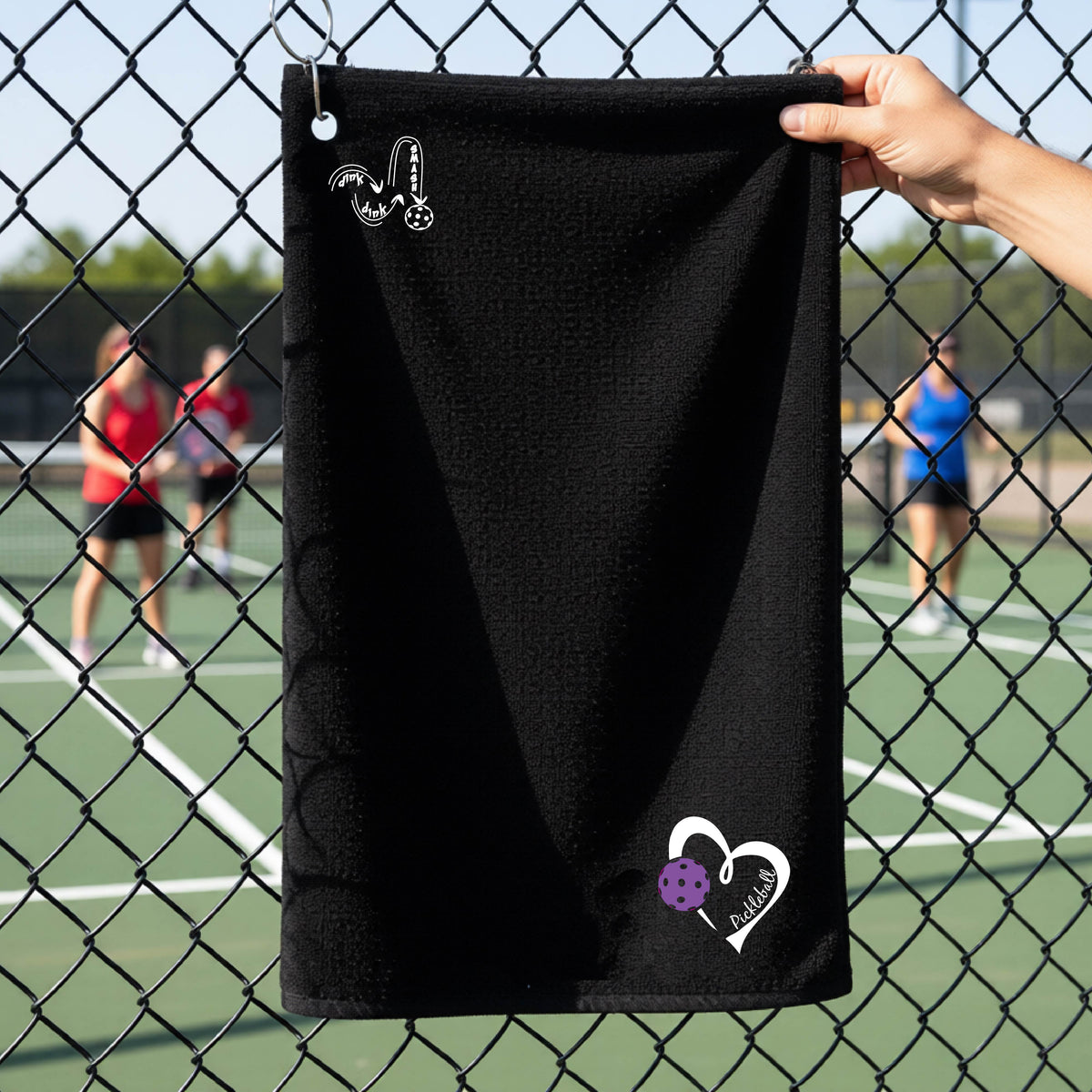 A black terry cloth towel held up by a hand in front of a chain-link fence and outdoor pickleball court. The towel has a white stylized logo near the top left 'DINK DINK SMASH' and a white heart logo with a purple pickleball graphic and the word 'Pickleball' near the bottom right.